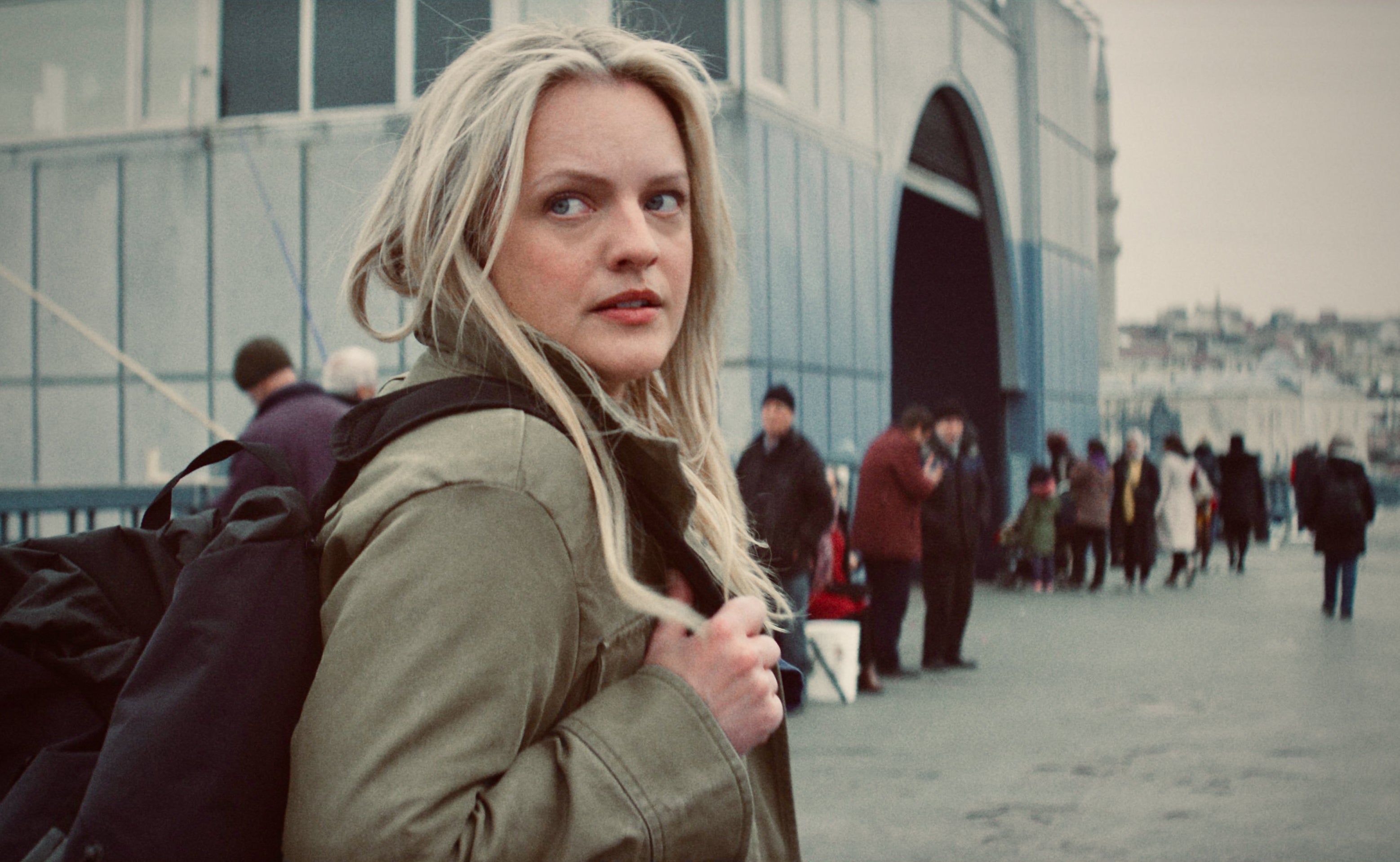 Elisabeth Moss in a scene, looking over shoulder with concerned expression, people in background in The Veil
