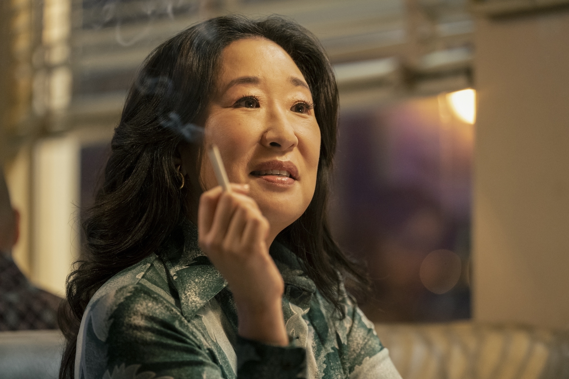 Sandra Oh wears a patterned blazer, holding a cigarette, with a contemplative expression in The Sympathizer