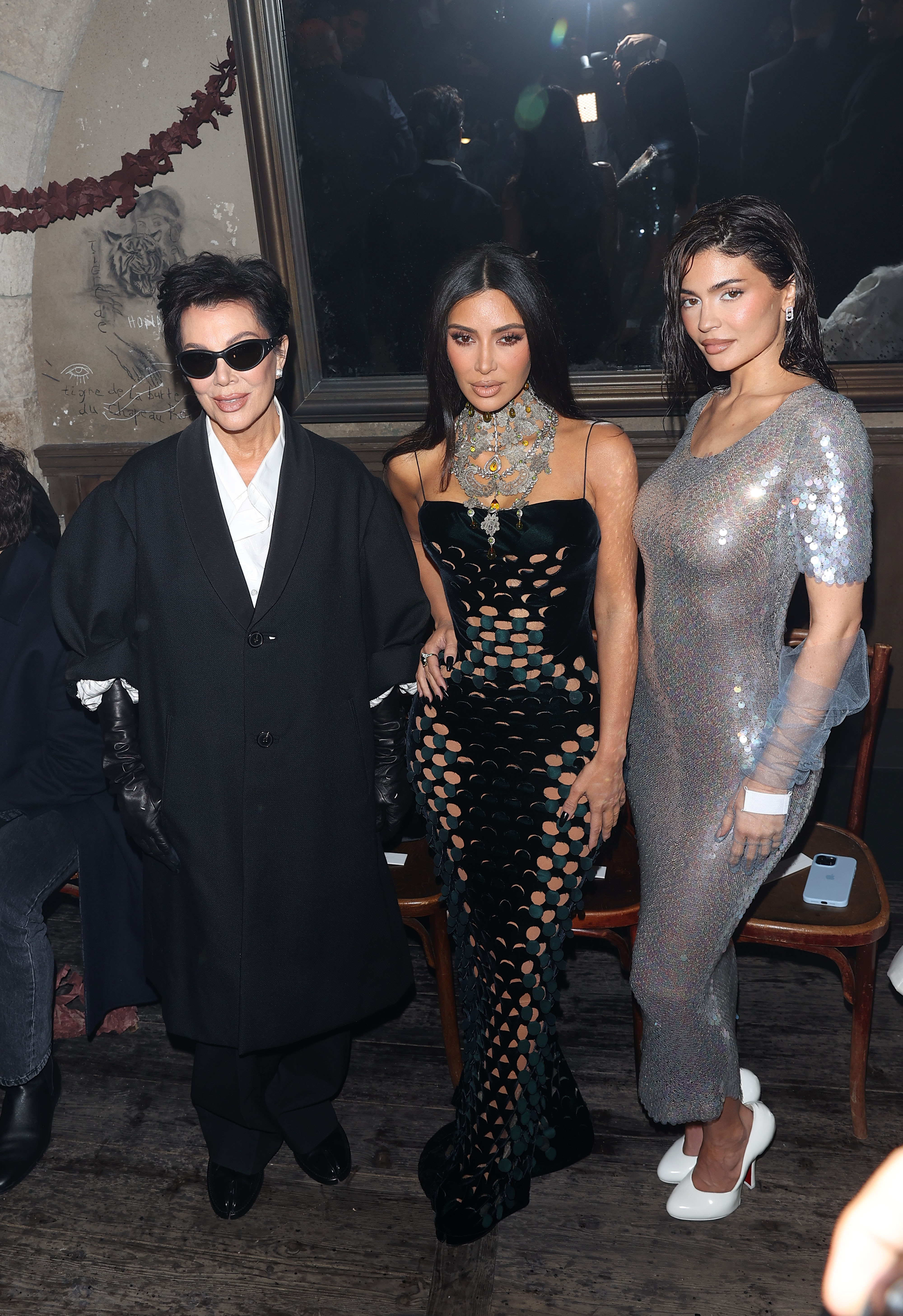 Kris Jenner, Kim Kardashian, and Kylie Jenner posing in stylish outfits at an event. Kim in a unique cut-out dress; Kylie in a shimmering gown