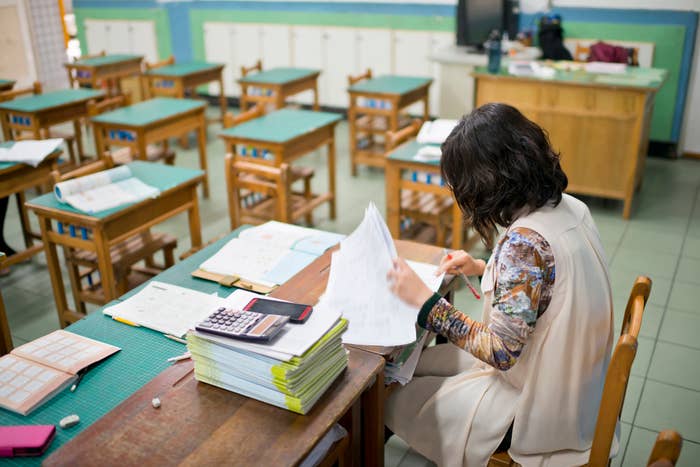 Teacher grading papers at a classroom desk, surrounded by stacked books and materials