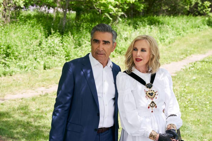 Eugene Levy and Catherine O&#x27;Hara stand outdoors, one in a suit and the other dressed in an outfit with a prominent heart design