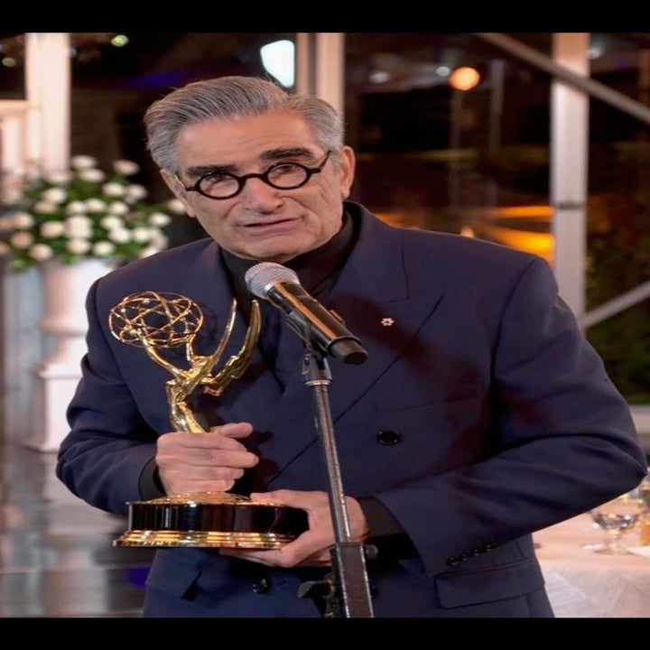 Man in glasses and suit holding an Emmy award, standing at a podium with audience in background