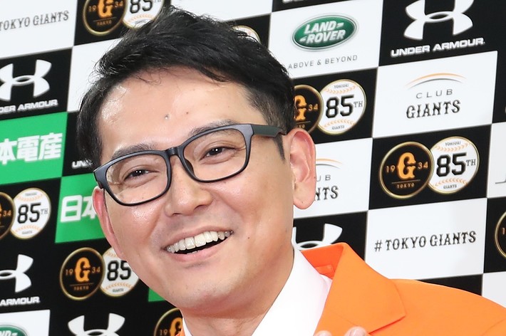 オレンジスーツを着た男性が笑顔でポーズをとっています。背景にはスポンサーロゴが並んでいます。