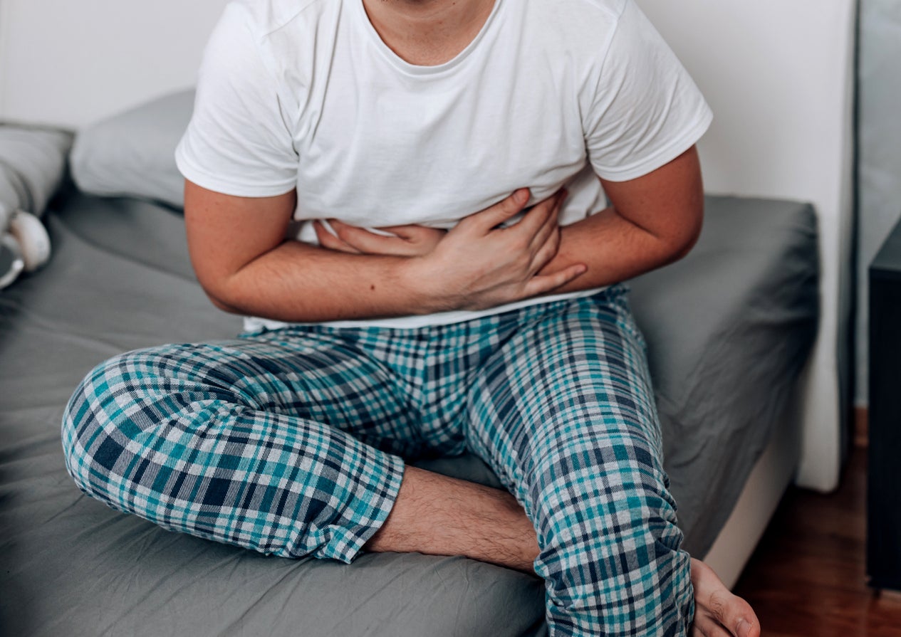 Person grimacing in pain, clutching stomach while sitting on the edge of a bed, wearing casual home attire
