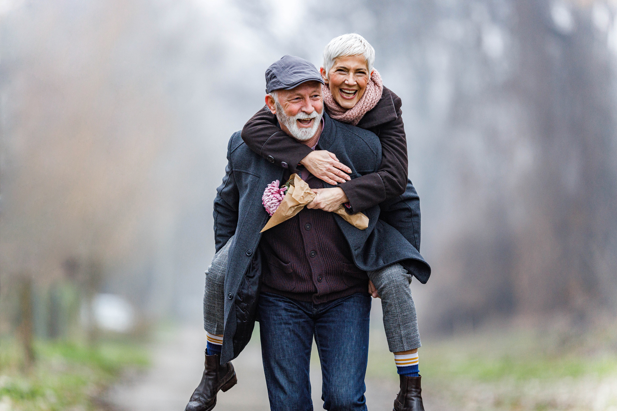 An older man gives a piggyback ride to a laughing woman, both dressed warmly for fall, sharing a joyful moment outdoors