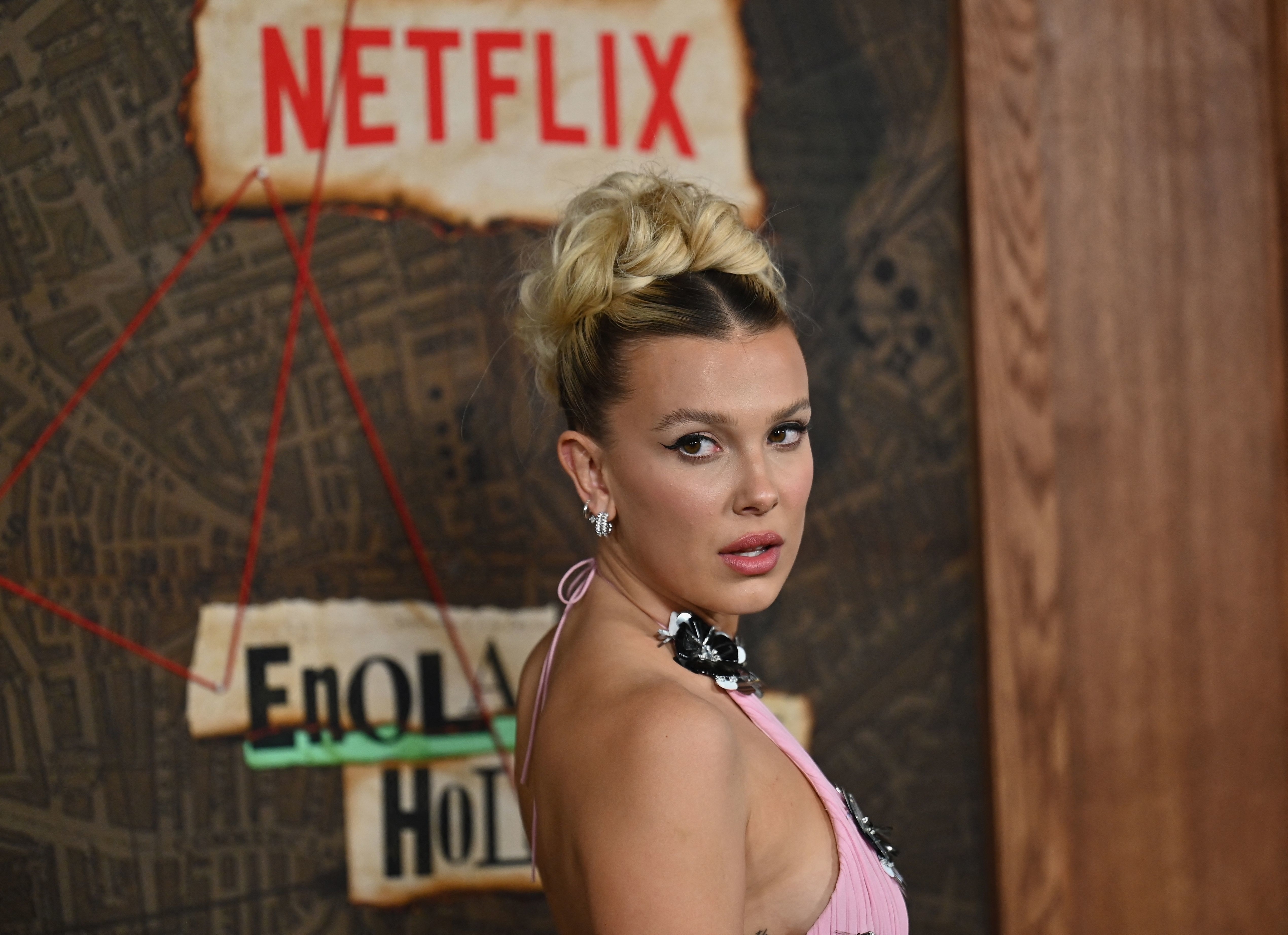 Millie in a dress and a statement necklace, with a bun hairstyle, on a red carpet with a Netflix logo background