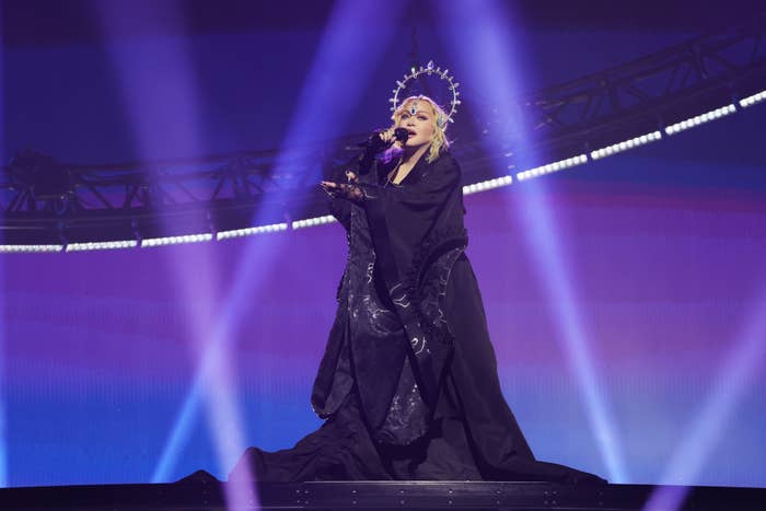 Performer in an embellished headpiece and black robe sings onstage with a microphone, backlit by stage lights