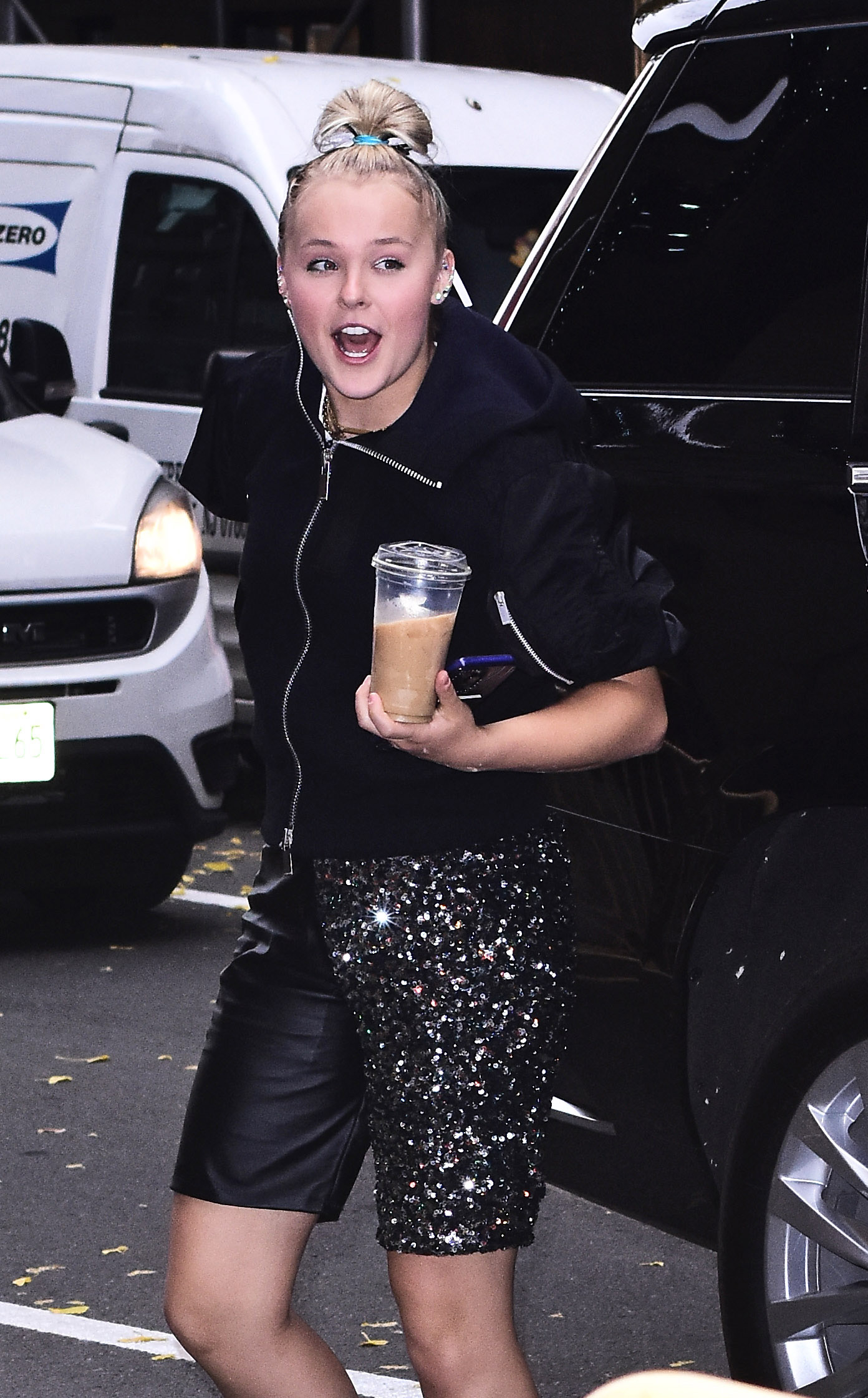 JoJo Siwa in a black jacket and sparkling skirt, exiting a vehicle with a beverage in hand