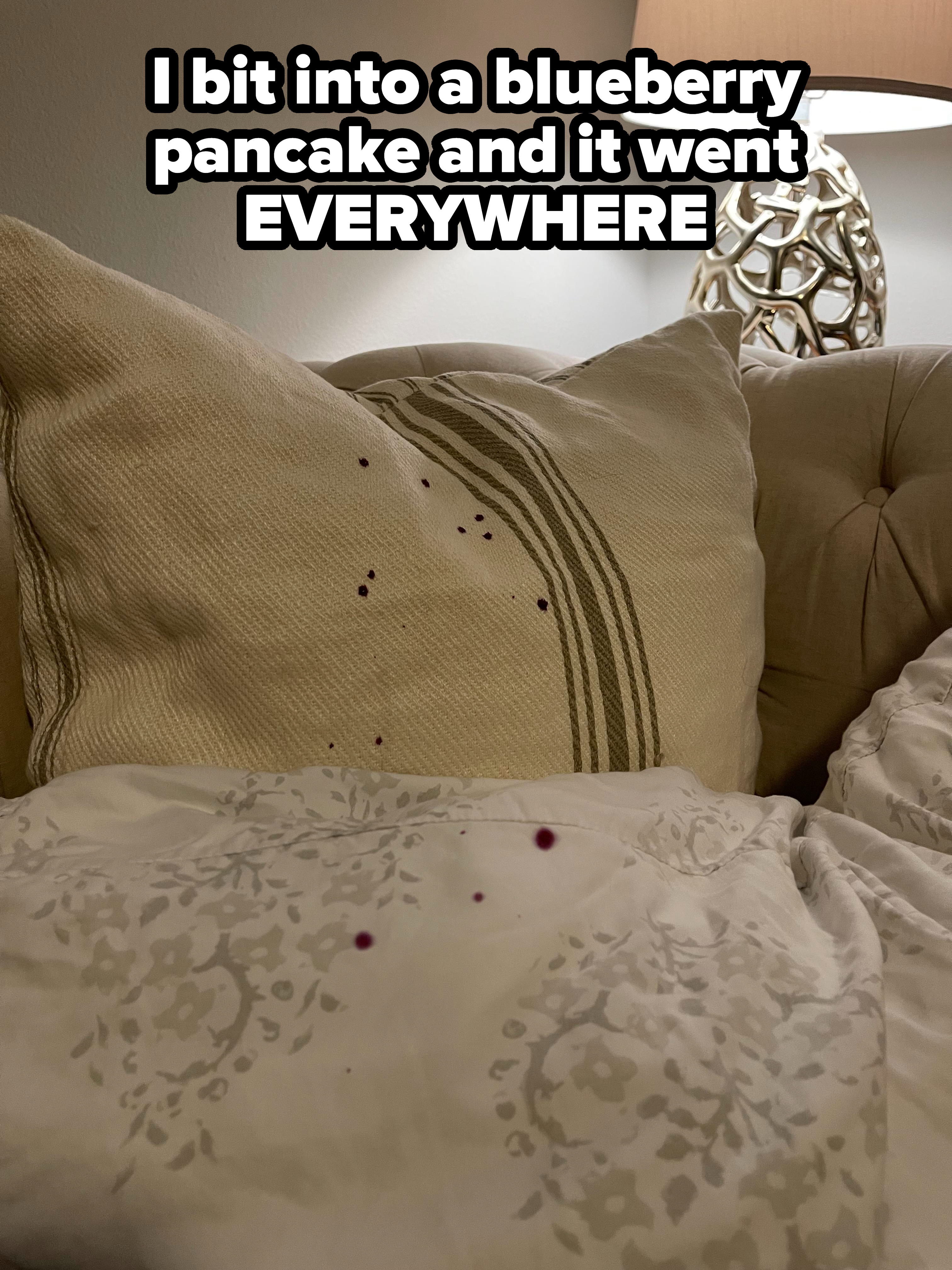 A close-up of a beige sofa with pillows and blueberry pieces/stains all over it