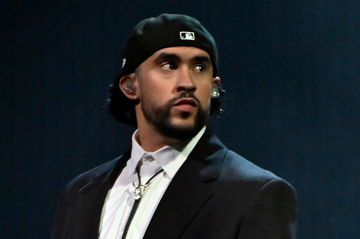 Rapper with a beanie and chain, wearing a jacket over a collared shirt, with a thoughtful expression