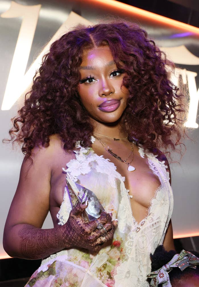 SZA posing, wearing a lace outfit with curly hair, holding an award