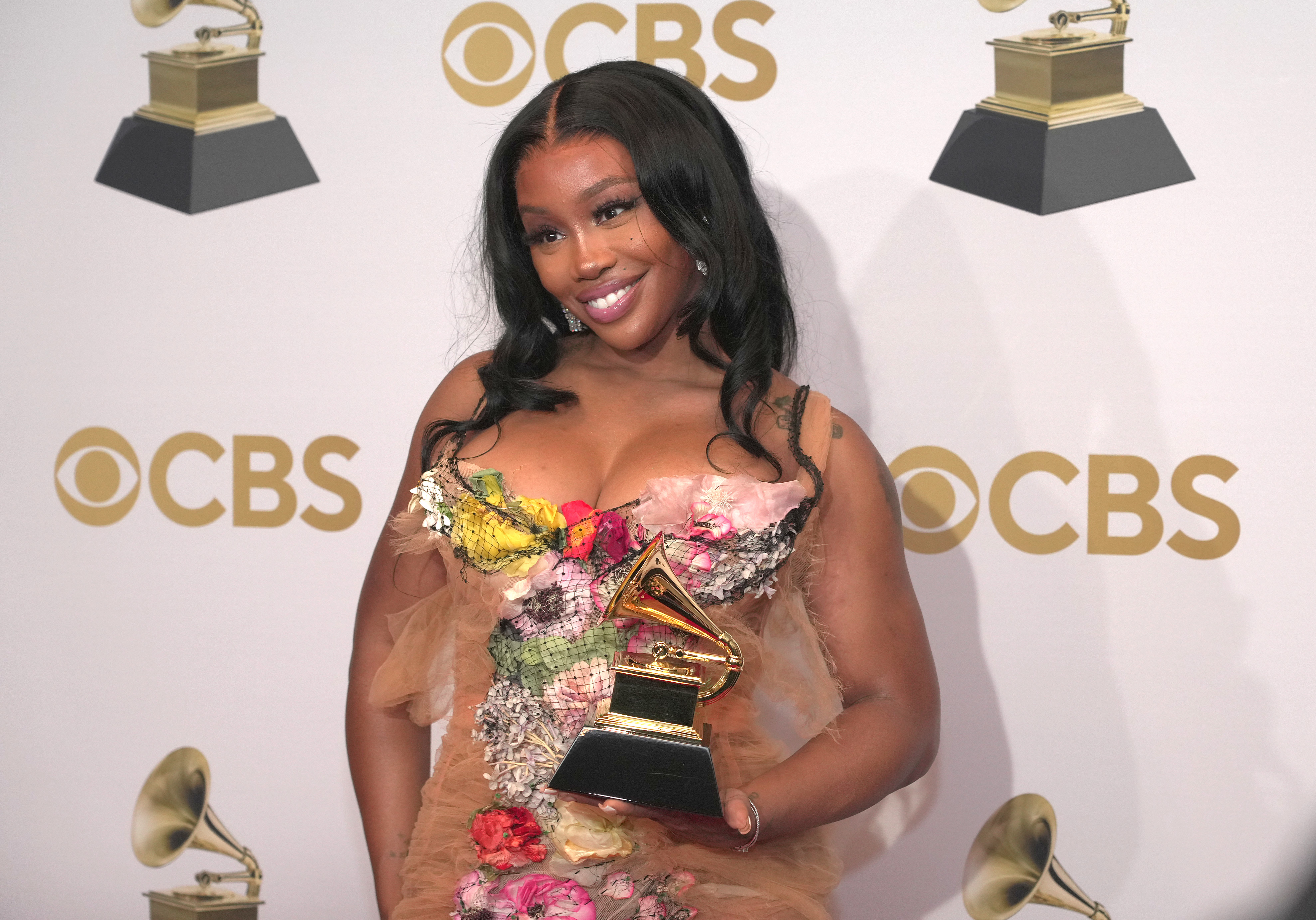 Sza posing with a Grammy Award, wearing a floral dress with sheer details