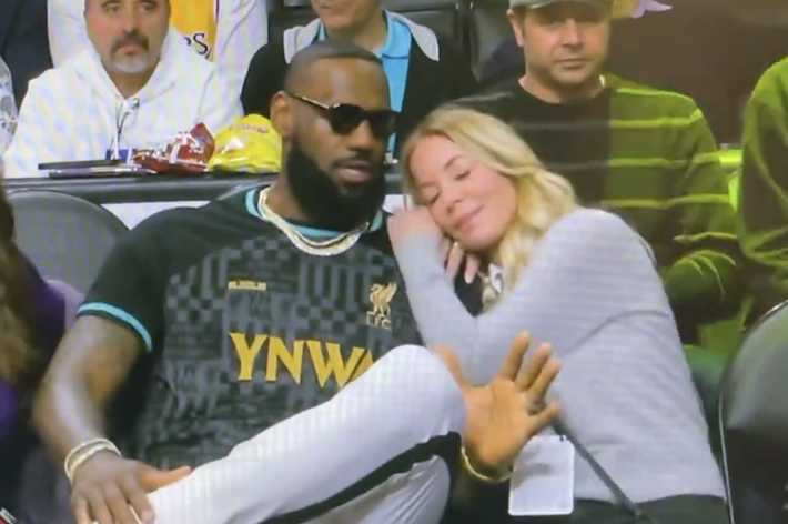 LeBron James in a black jersey seated courtside next to a woman in a grey top