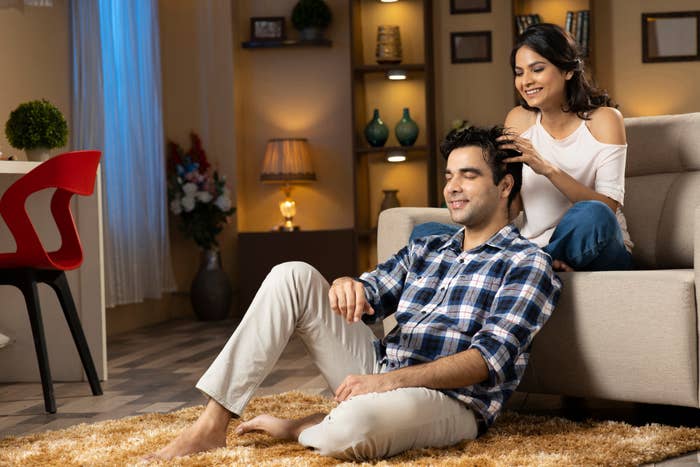 A couple relaxing at home, with the woman affectionately styling the man&#x27;s hair as they smile
