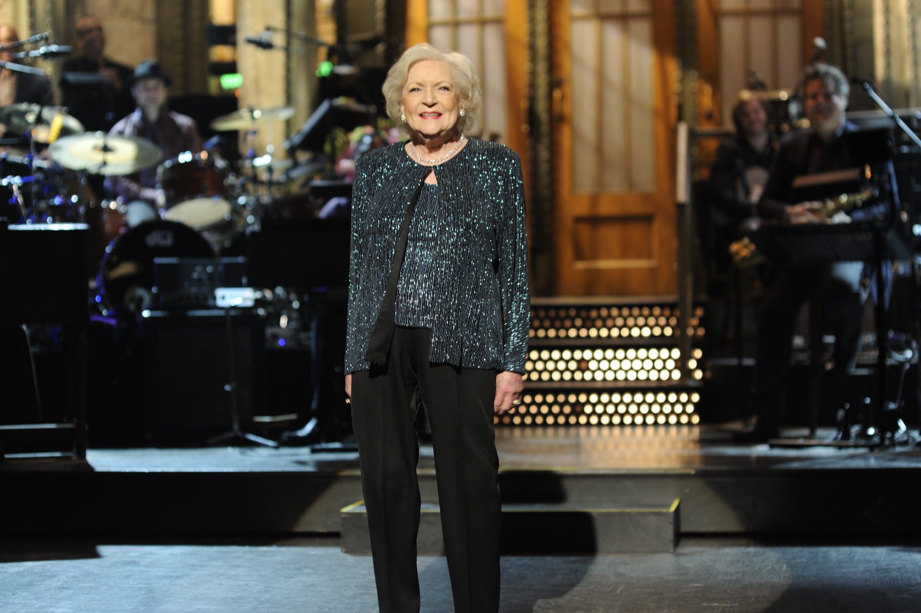 Betty White stands smiling on stage in a glittery jacket with a band in the background