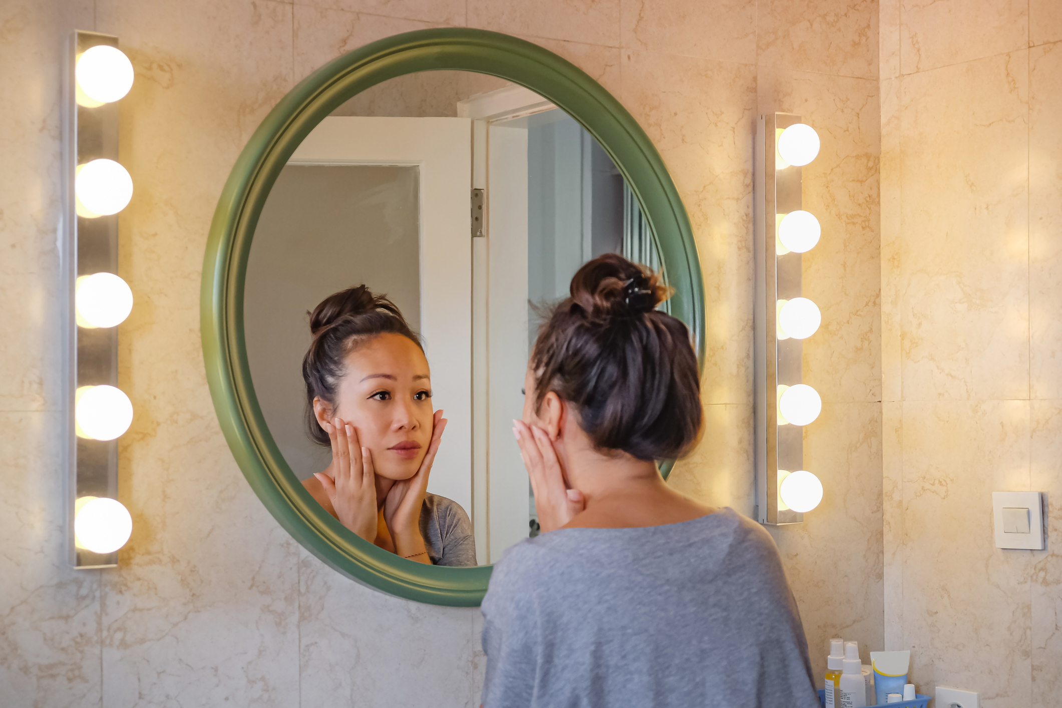 Woman examining her face in a lit bathroom mirror, with a focus on skincare