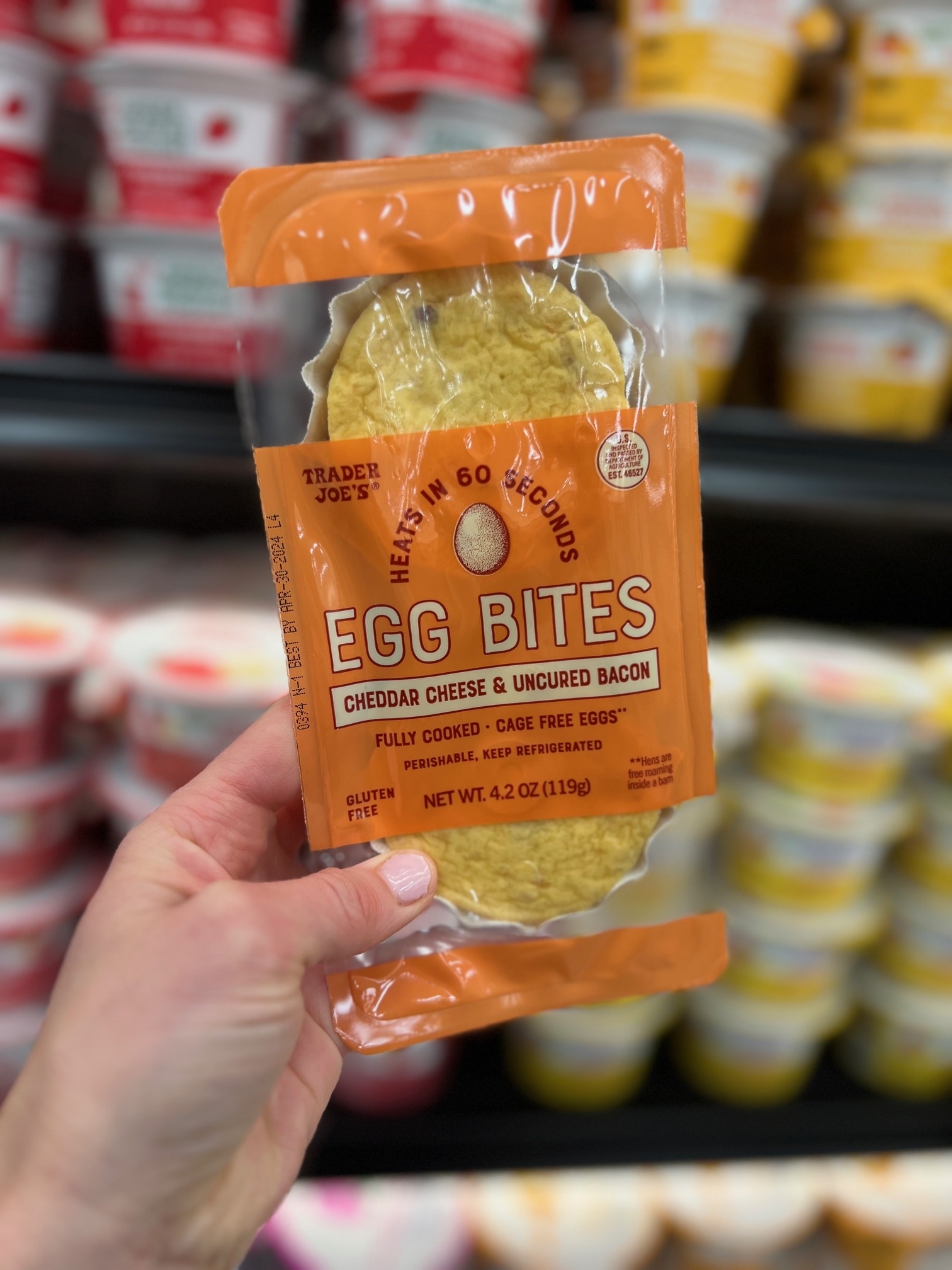Hand holding a package of Trader Joe&#x27;s Egg Bites with Cheddar Cheese &amp;amp; Uncured Bacon against store shelves