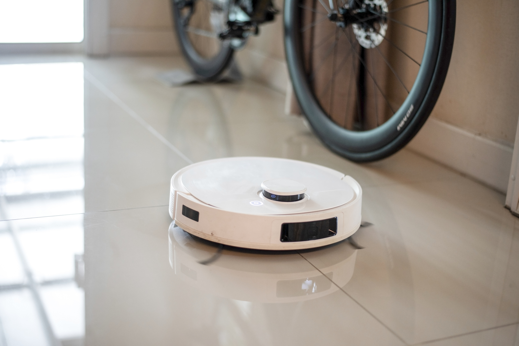 A robotic vacuum cleans the tiled floor with a bike mounted on the wall in the background