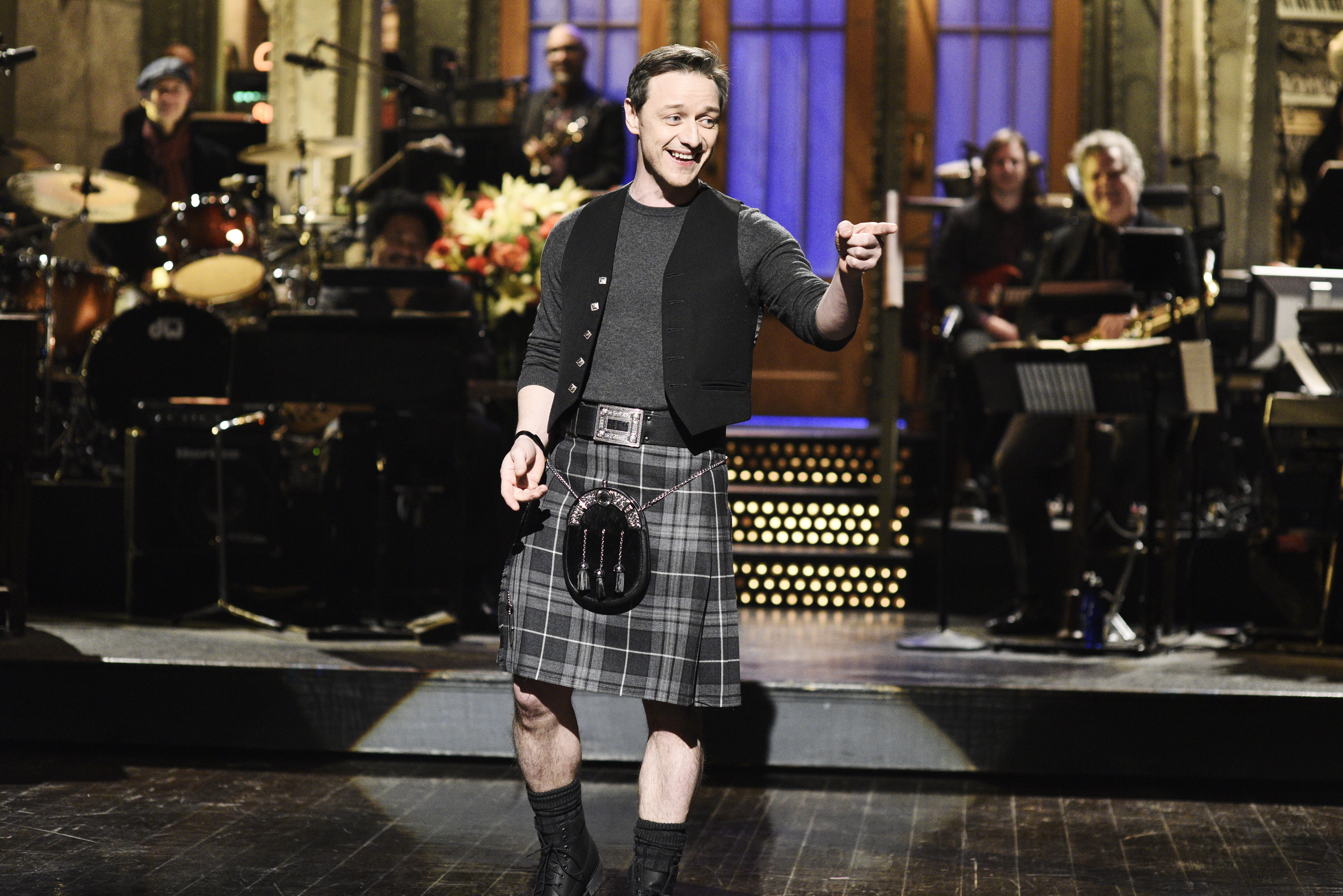 Person in a kilt and vest smiling and gesturing on a stage with a band in the background