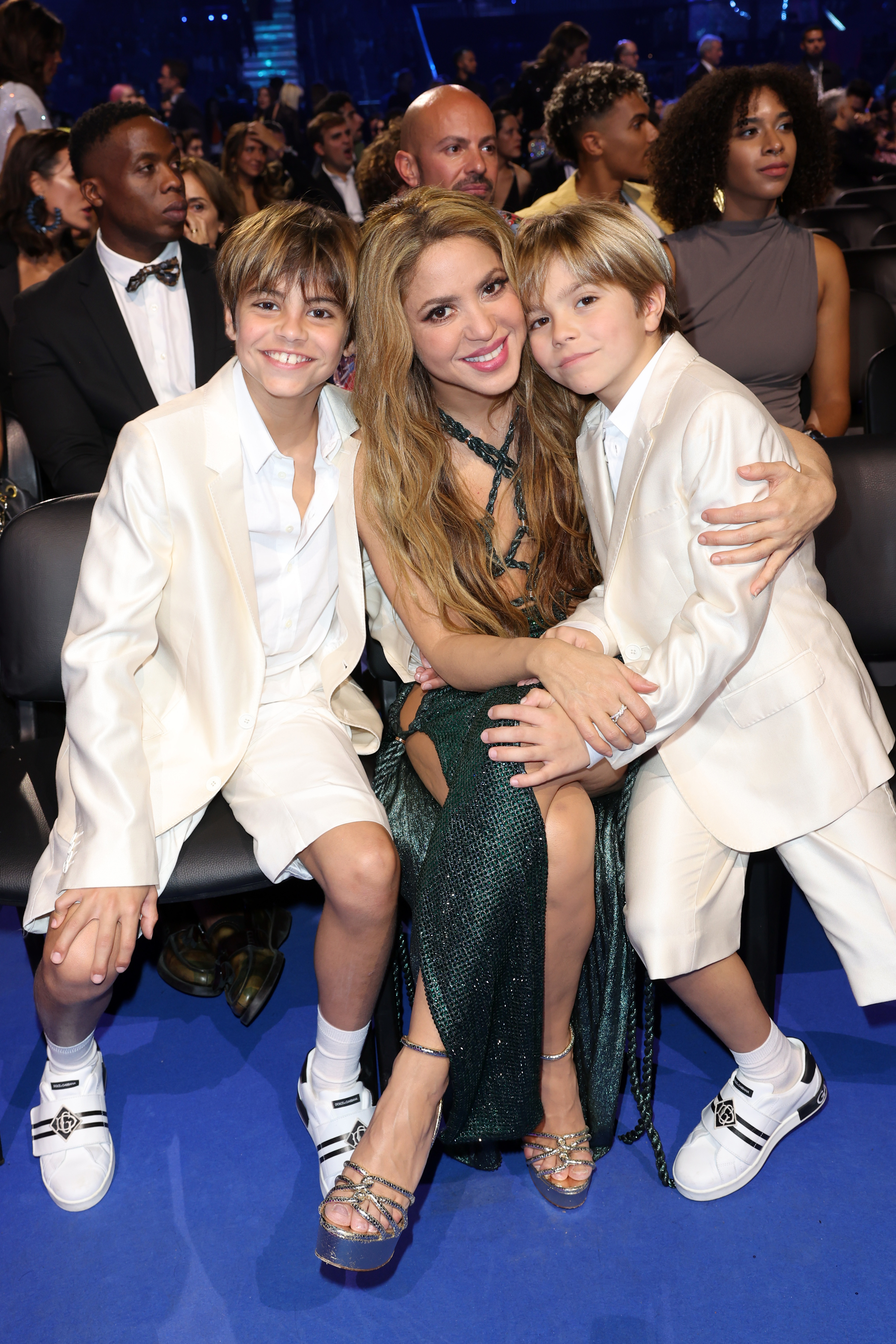 Shakira seated with her two boys in formal wear at an event, all smiling for the camera