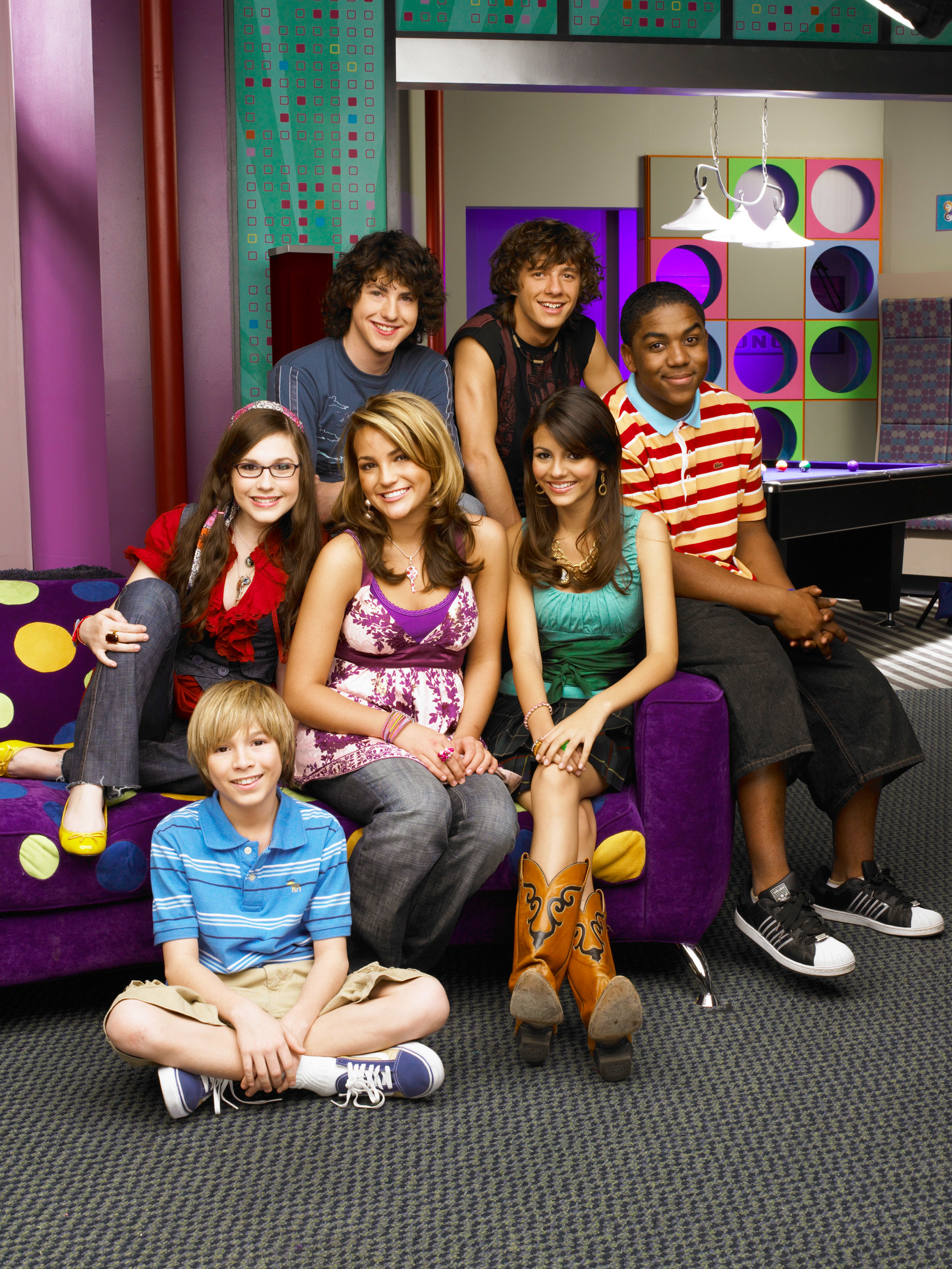 Seven cast members of Zoey 101 posing together on a set with colorful backgrounds and modern furniture