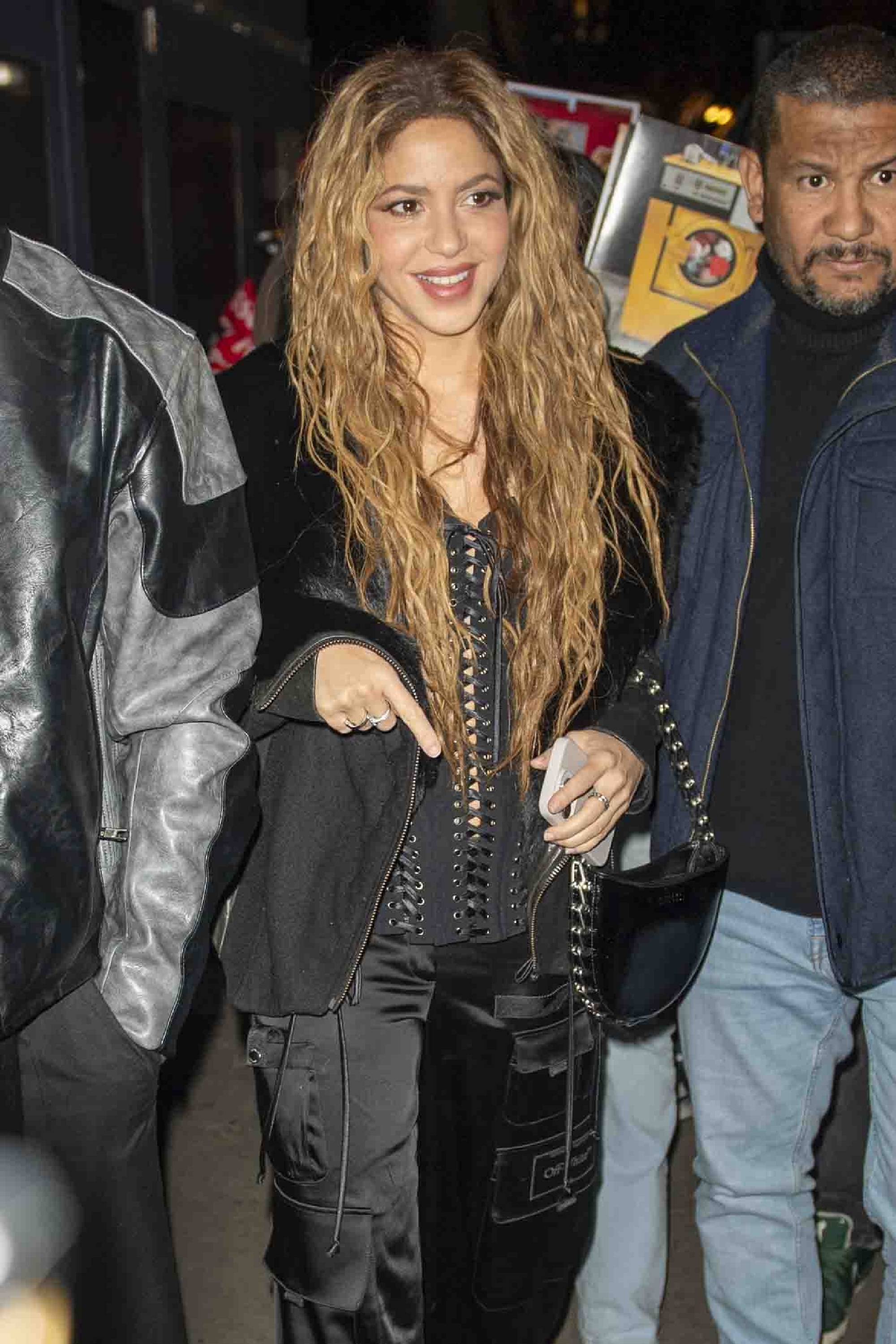 Shakira smiles in a laced outfit and carries a handbag, accompanied by a man on a street at night