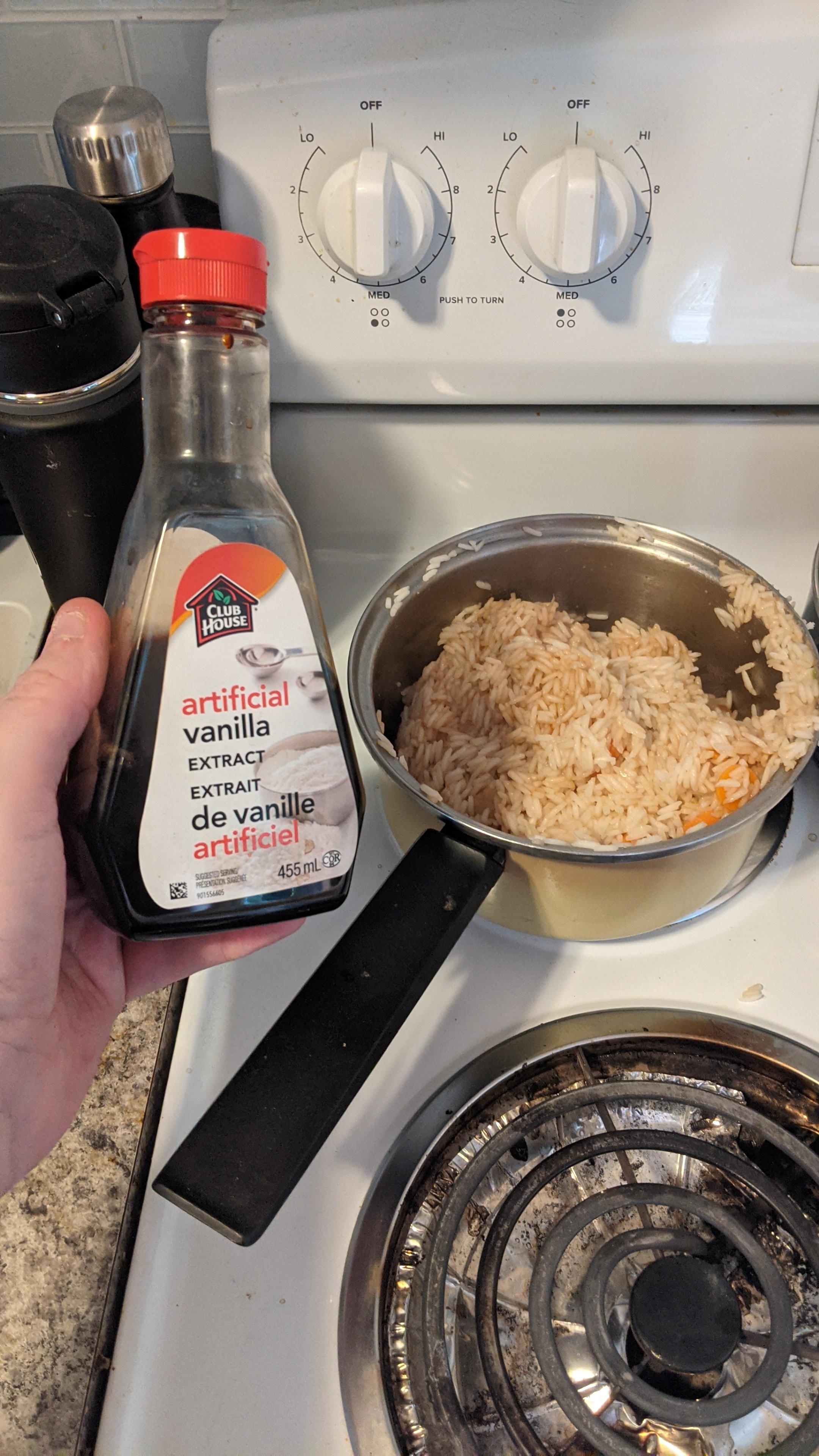 Person holding a bottle of vanilla extract next to a pot of rice on a stove