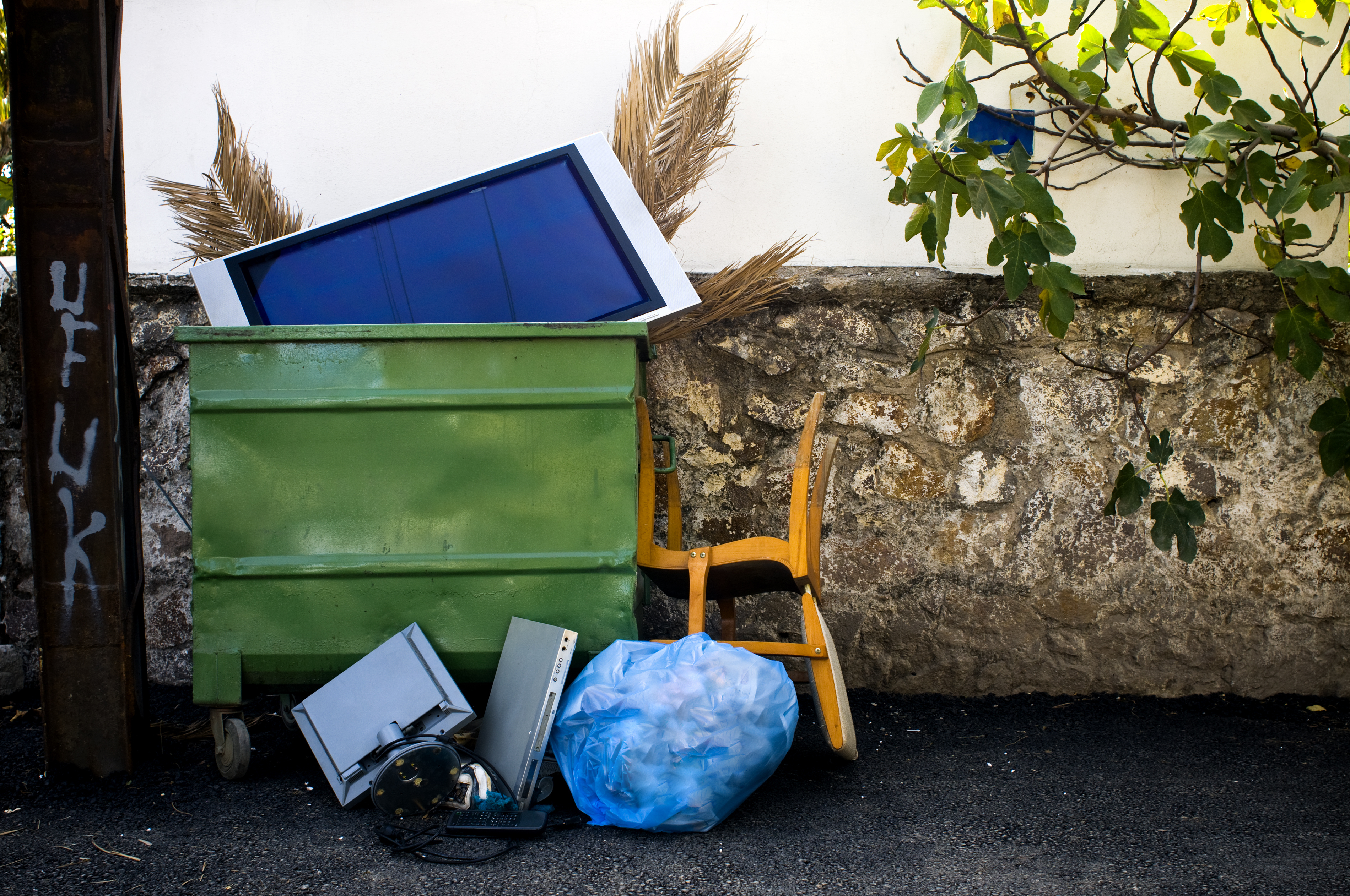 Overturned green dumpster with electronic waste and a blue bag, next to a wall and under a tree