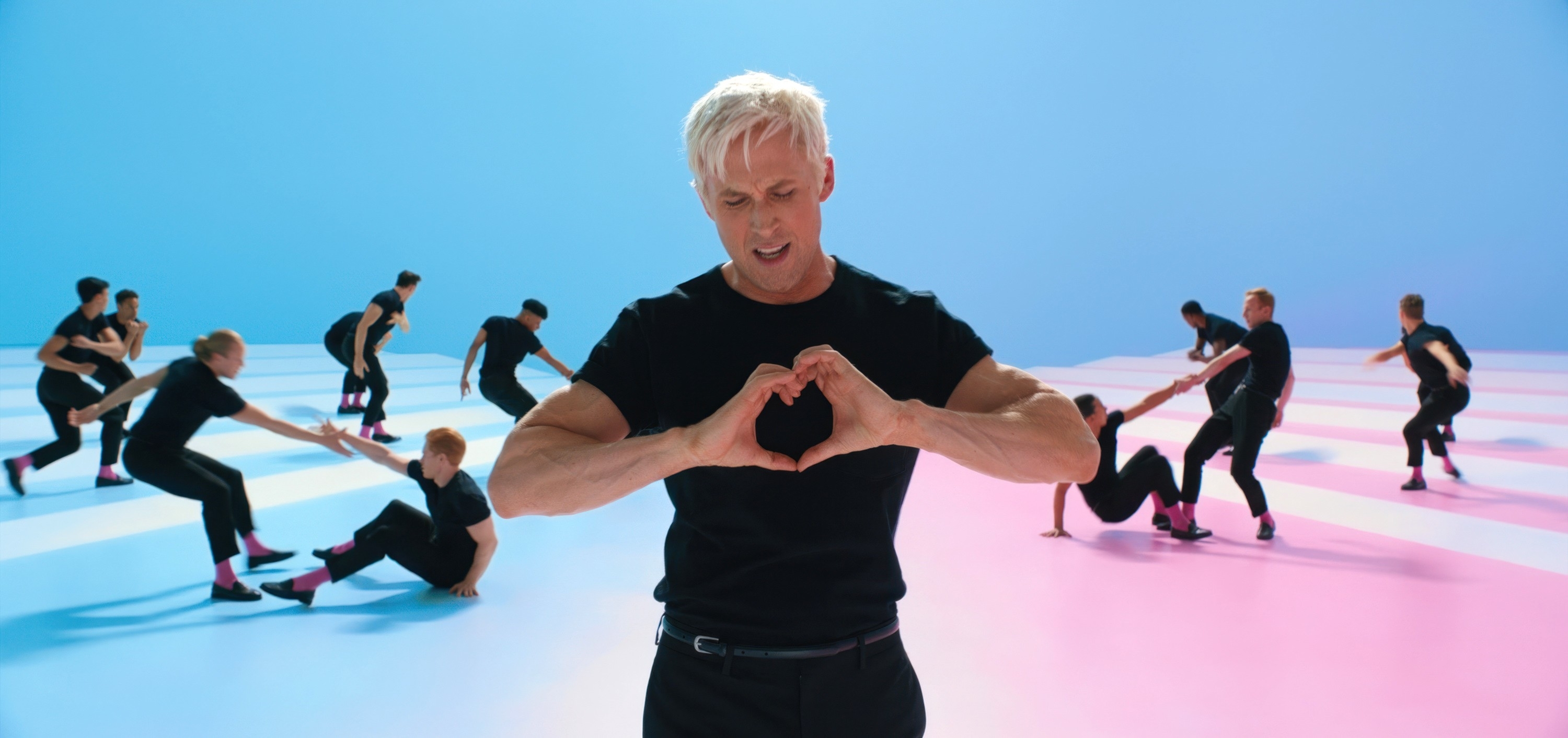 Ryan Gosling as Ken making a heart shape with his hands, surrounded by dancers in matching outfits, on a vibrant stage
