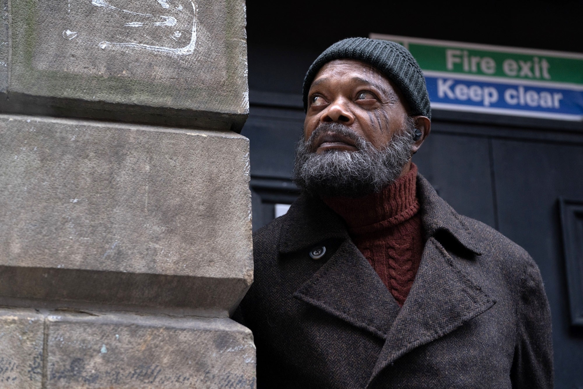 Nick Fury with a beard and a knit cap, wearing a layered outfit, looking upwards beside a building with a sign