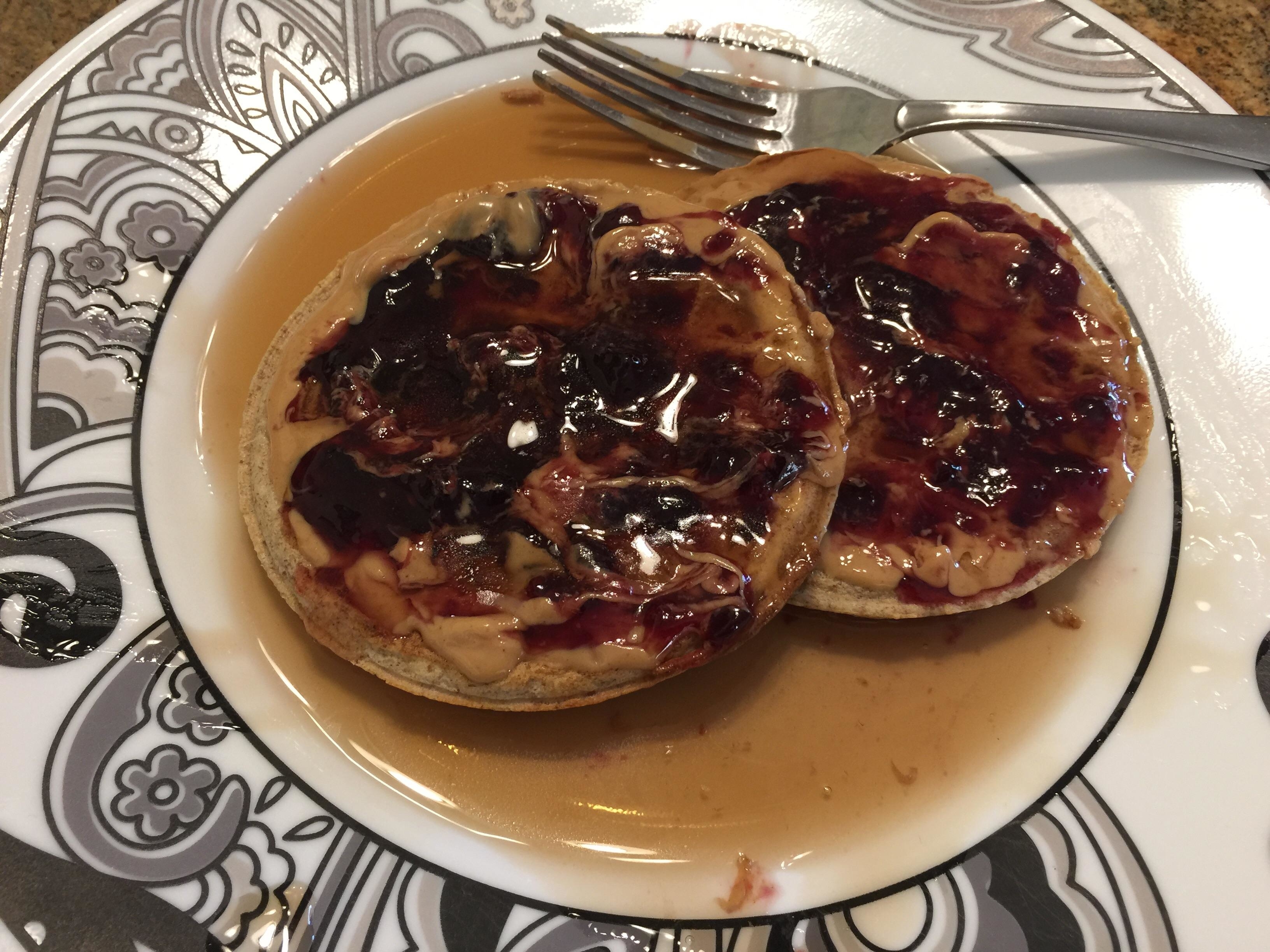 Two pancakes on a plate with syrup and jelly, alongside a fork