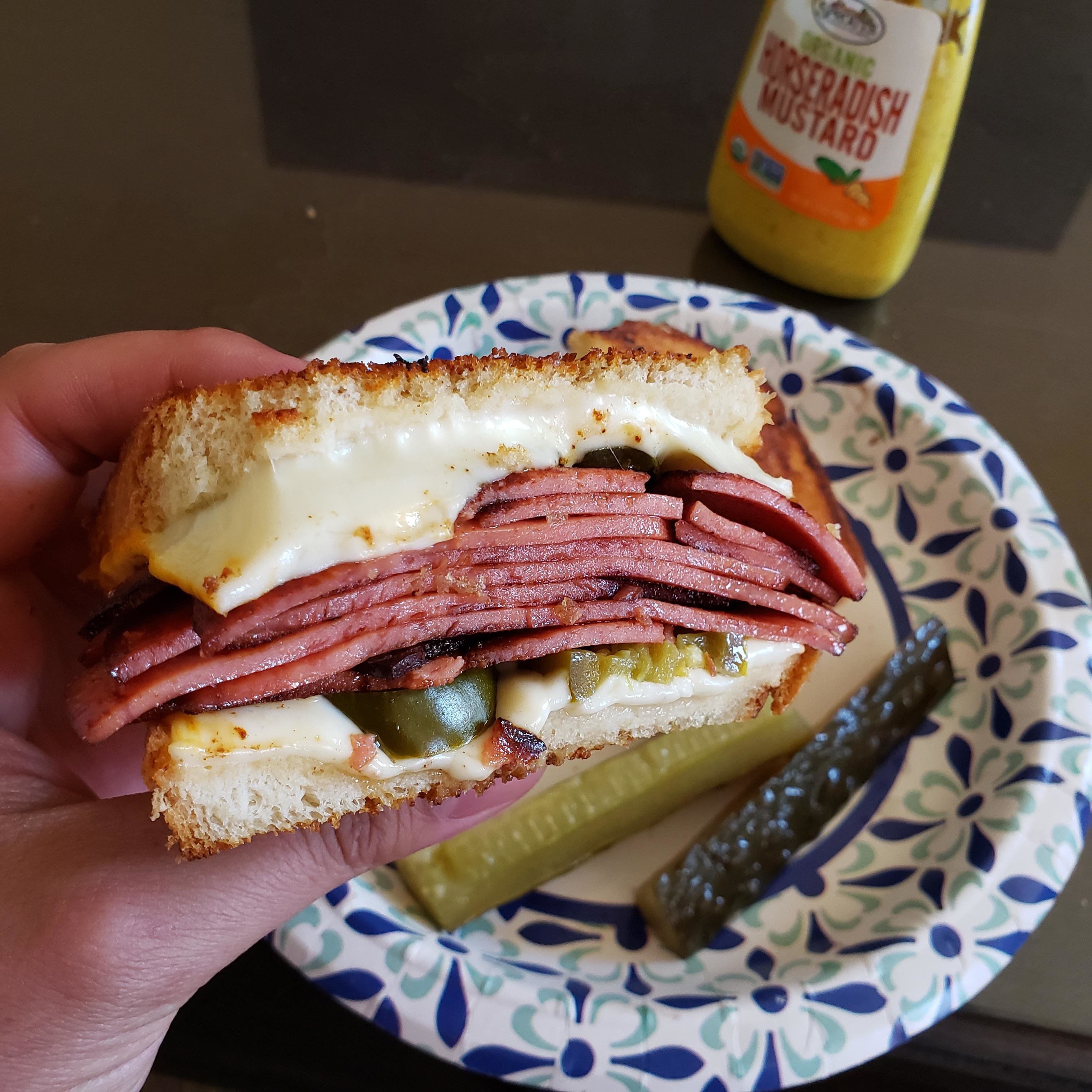 Hand holding a sandwich with multiple salami slices, cheese, and pickles, with a whole pickle on the side