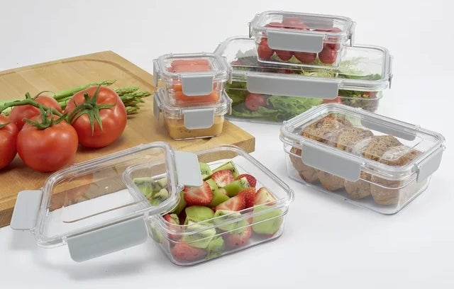 Assorted clear food storage containers with fresh produce and sandwiches inside, displayed on a table with tomatoes and a cutting board nearby