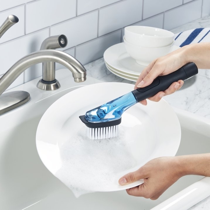 Person washing a dish with a handheld soap-dispensing brush in a kitchen sink