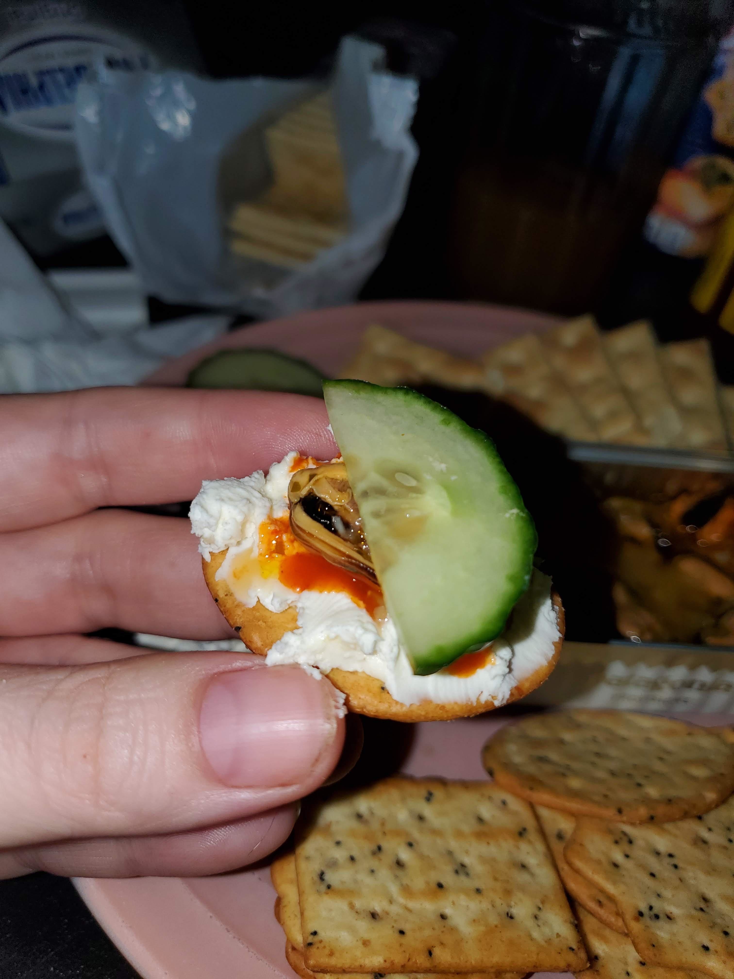 Hand holding cracker with cream cheese, a slice of cucumber, and a mussel, with more crackers in background