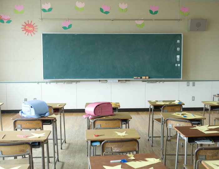 Empty classroom with desks, backpacks, and papers, chalkboard in front. No individuals present