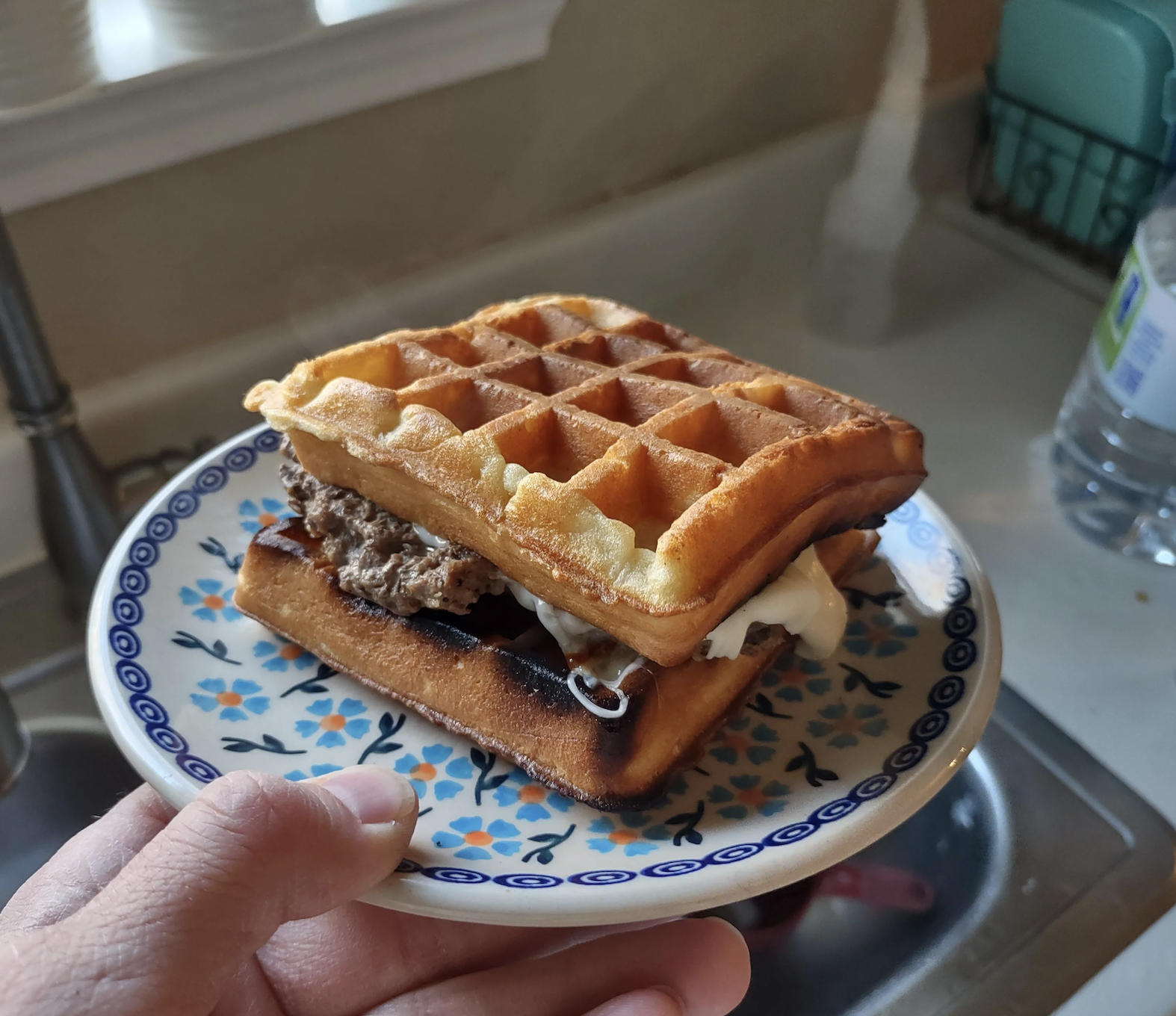 A person holding a plate with a waffle sandwich containing eggs and sausage