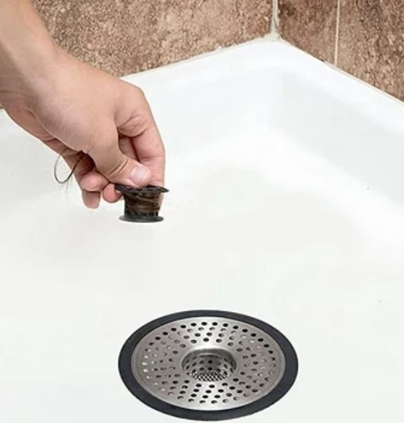 Hand removing hair from a bathtub drain next to a drain strainer