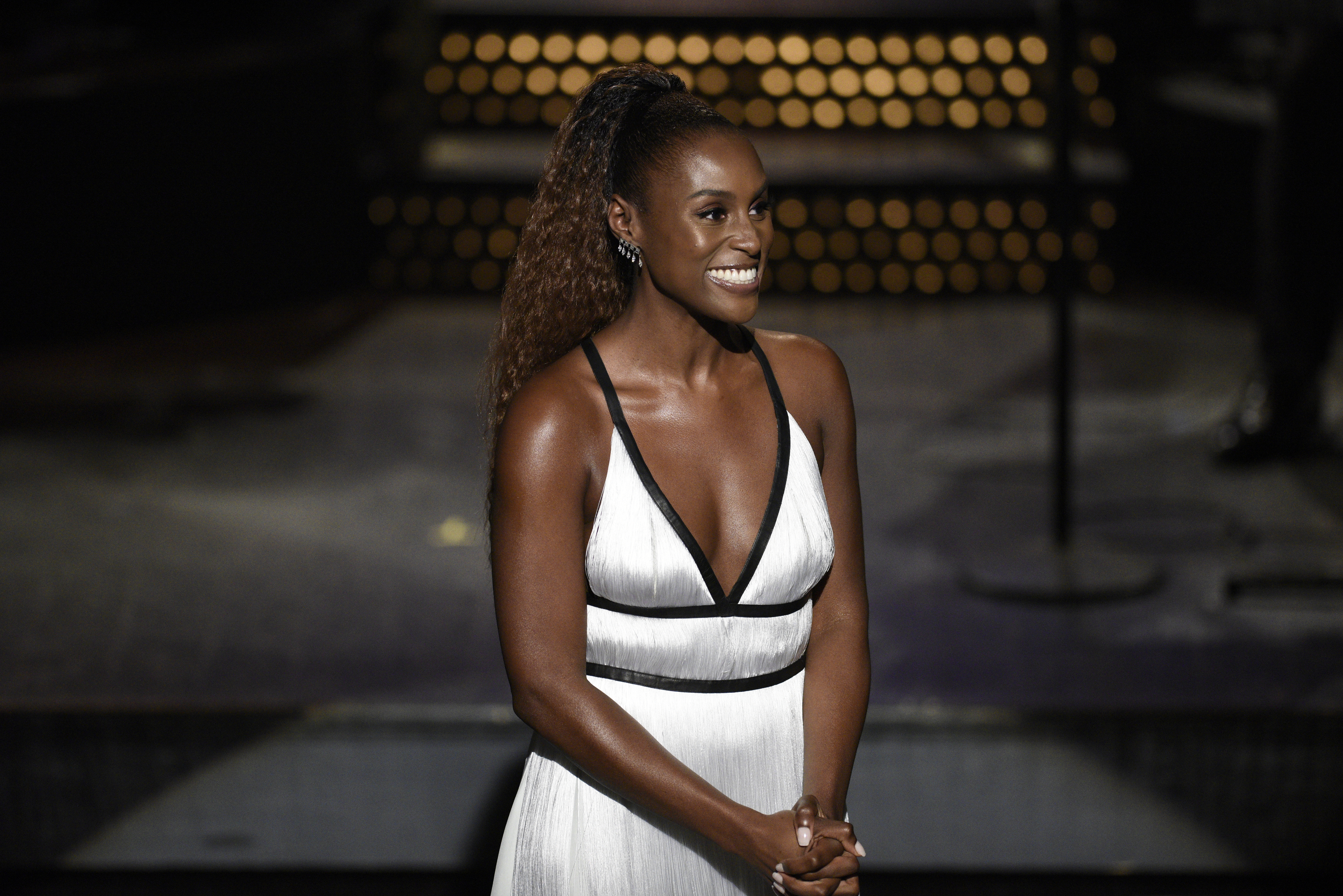 Actress in white V-neck dress smiles on stage, hands clasped together