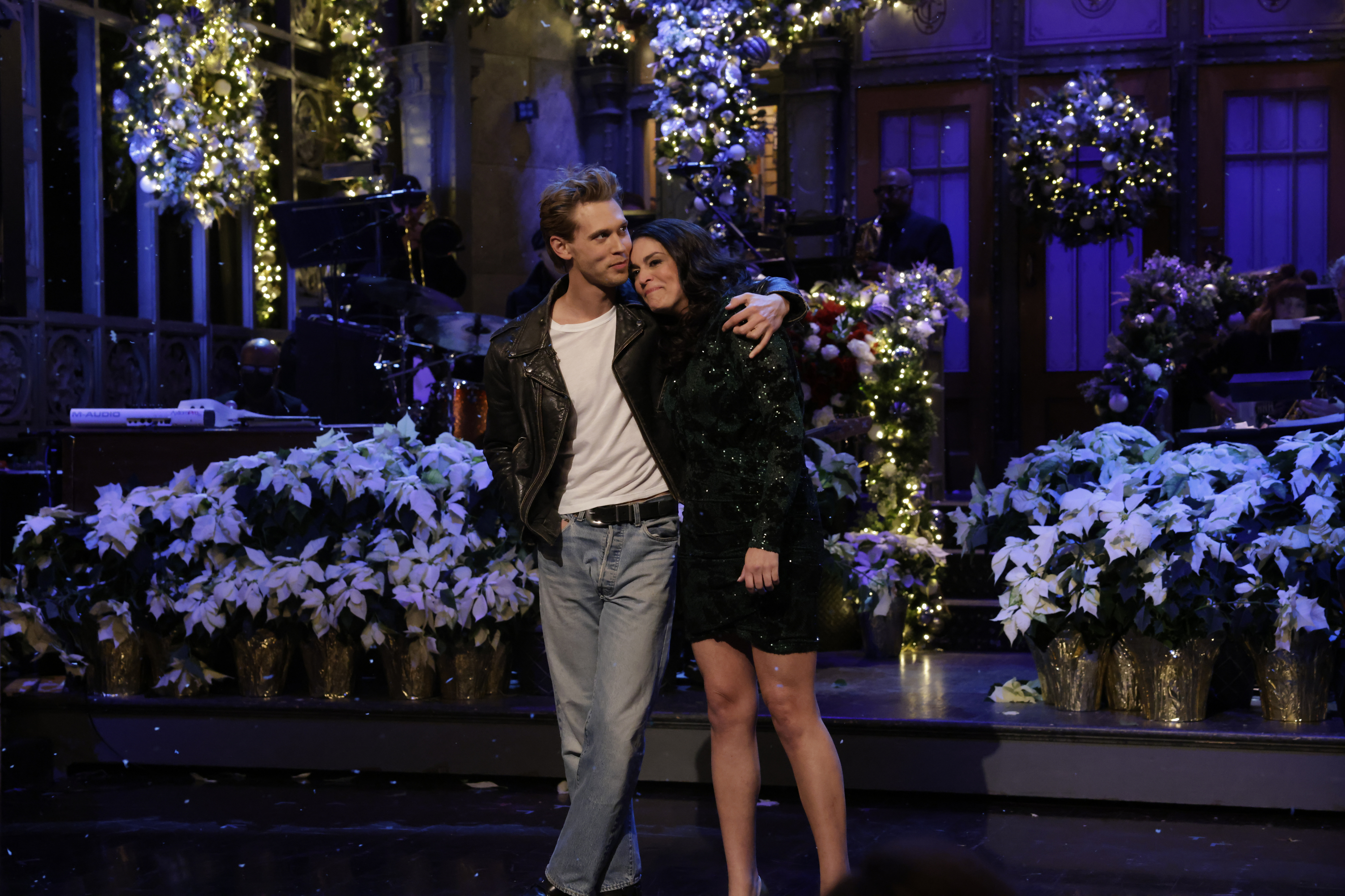 Two actors on SNL stage with holiday decor, one in leather jacket and jeans, the other in a feathered dress