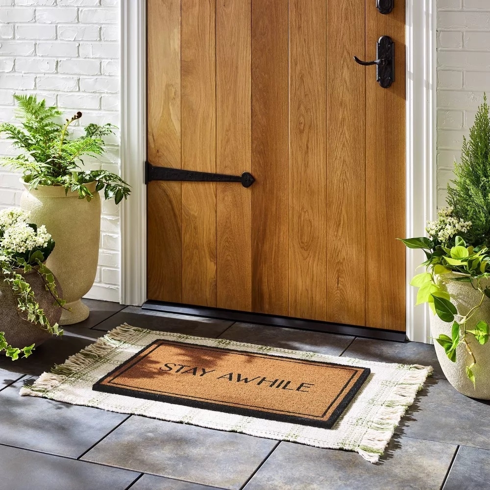 Door mat with &quot;STAY AWHILE&quot; on it, framed by plants, positioned at a wooden front door entrance