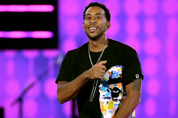 Ludacris wearing a graphic tee and jeans, standing with a mic in front of a stage background