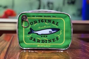 sardine can-shaped bag on a wooden surface, with a fish illustration and the text "ORIGINAL Sardines."