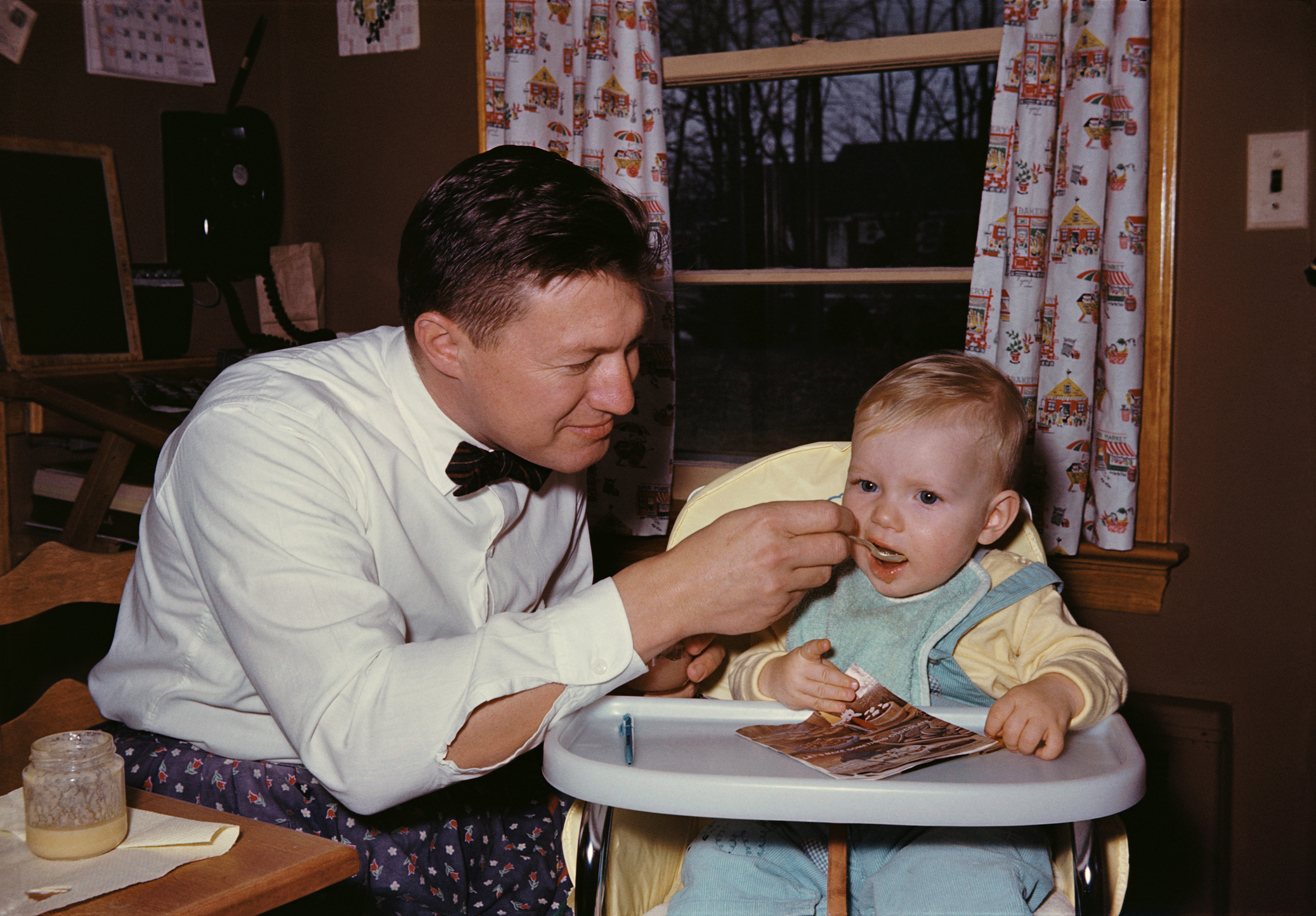 Man feeding a child in a high chair; both are indoors with vintage decor
