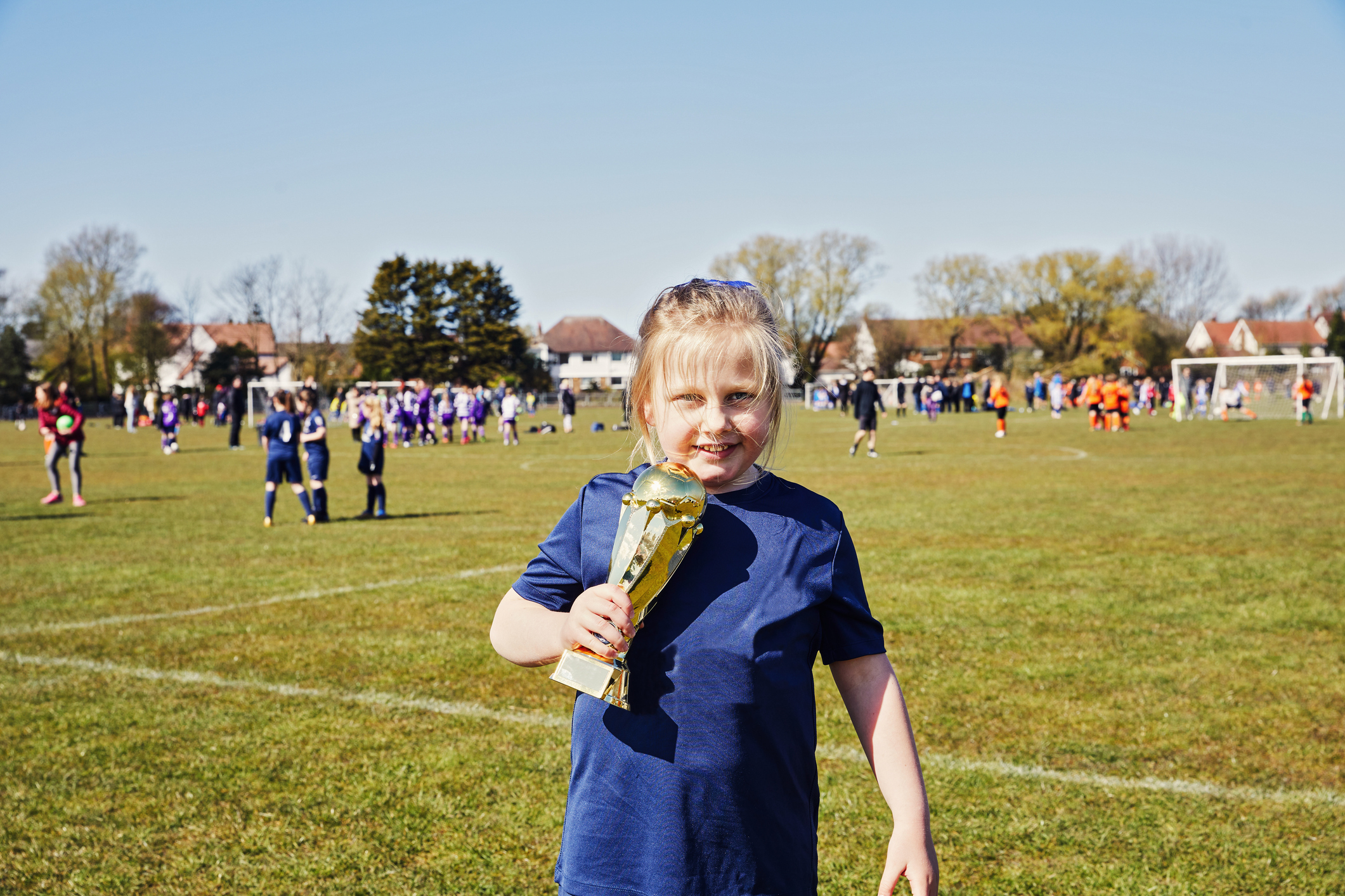 Child holding a trophy on a soccer field with players in the background