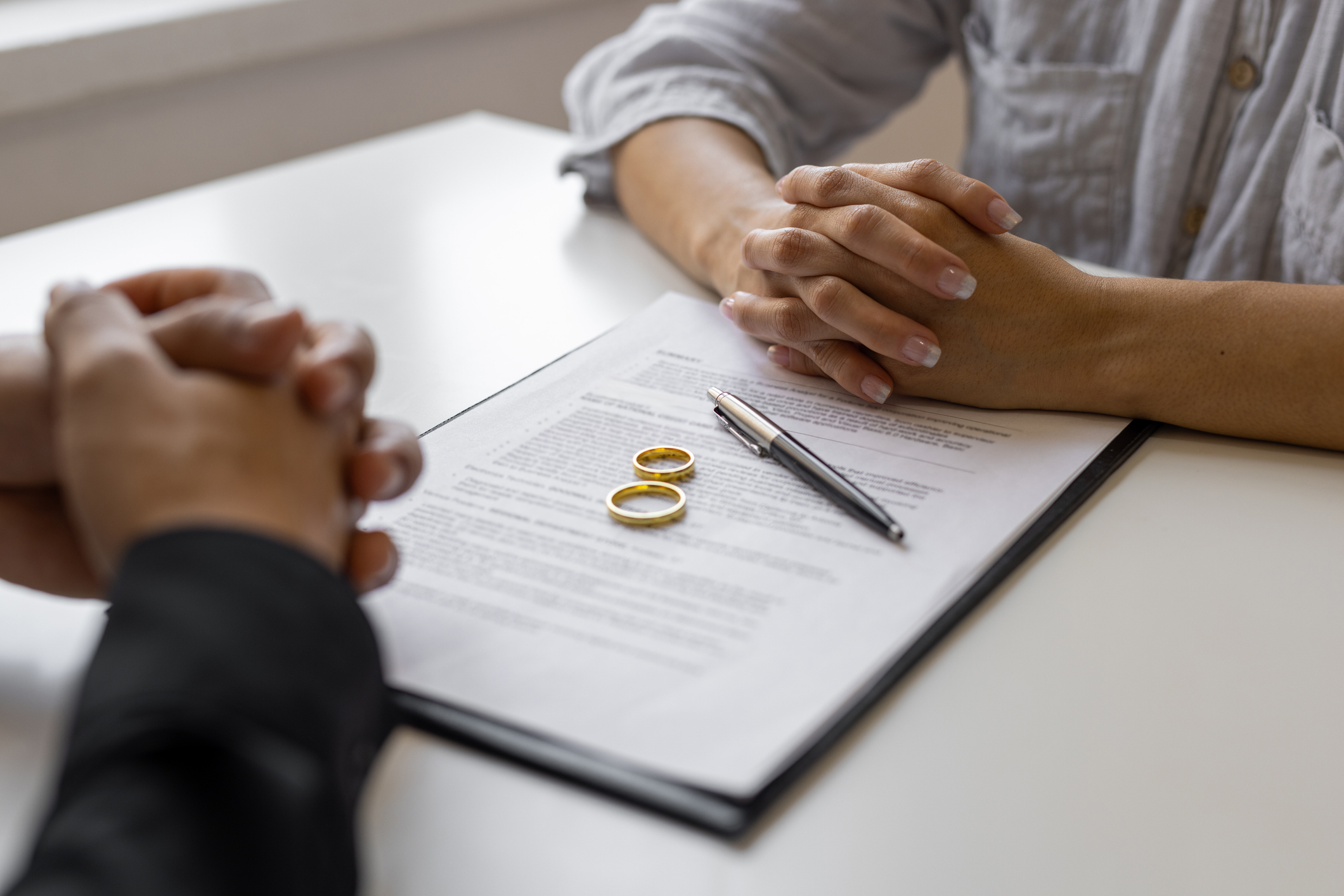 Two people sitting across from each other with their hands folded near wedding rings and documents on a table