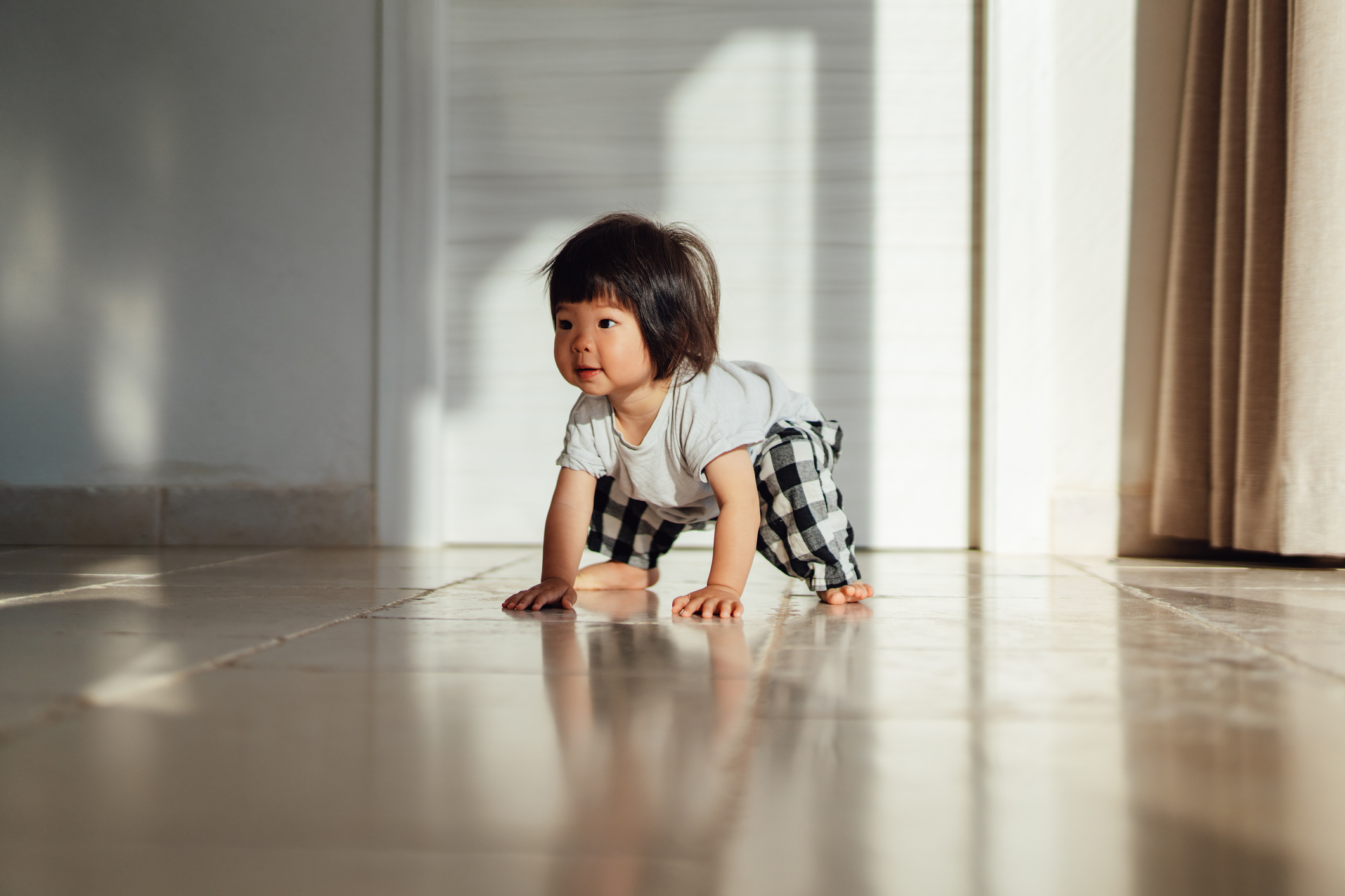 Toddler crouching on a tiled floor with sunlight streaming in, looking to the side