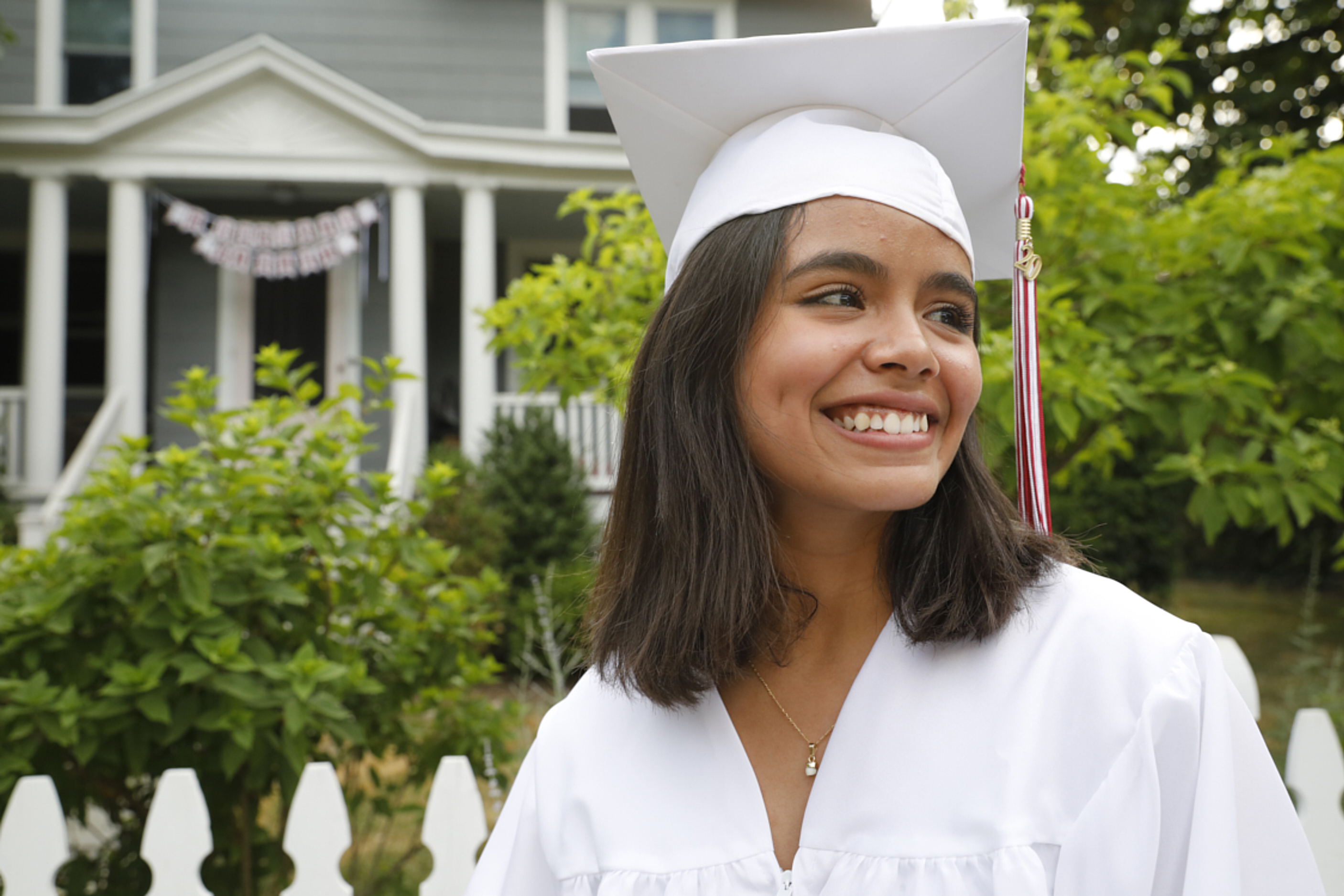 Graduate in white cap and gown smiling outdoors with a house in the background