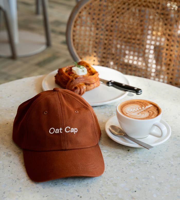 Embroidered &quot;Oat Cap&quot; hat on a table beside a pastry and a cup of cappuccino in a cafe setting