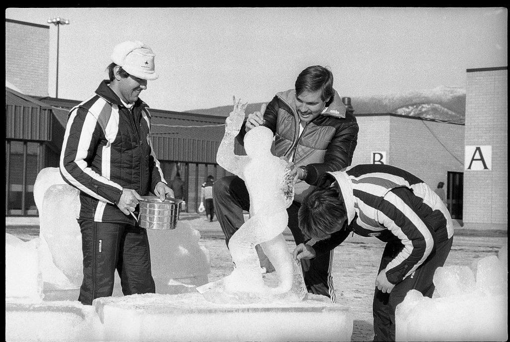 Three people carving an ice sculpture outdoors near a building with the letter &#x27;A&#x27; on it